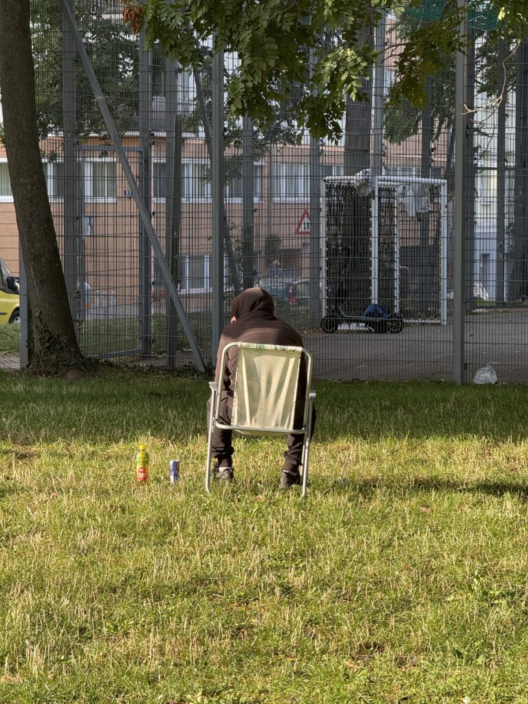 Mitten in der Wiese - vor einem Fußballkäfig - sitzt auf einem Klappstuhl ein Mann mit schwarzen Kapuzenpulli. Die Kapuze ist über den Kopf gezogen.
