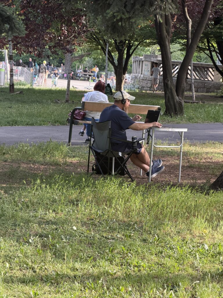Eine Person sitzt in einem Park auf einem Klappstuhl an einem kleinen Tisch und arbeitet an einem Laptop. Sie trägt Kopfhörer und eine helle Kappe.