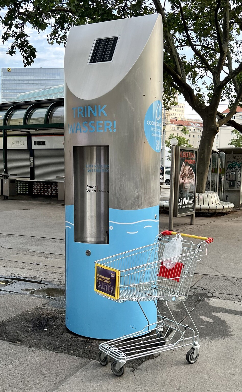 Öffentlicher Platz. Neben einem Wasserspender steht ein Einkaufswagen.