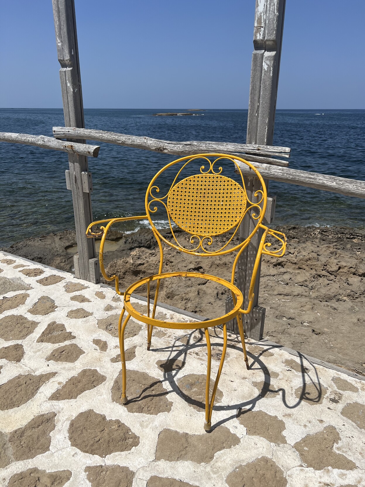 Meeresstrand. Davor Pflaster mit einigen Holzpfählen Richtung Sand. Auf dem Pflaster steht ein gelber Metallstuhl dem die Sitzfläche fehlt. 