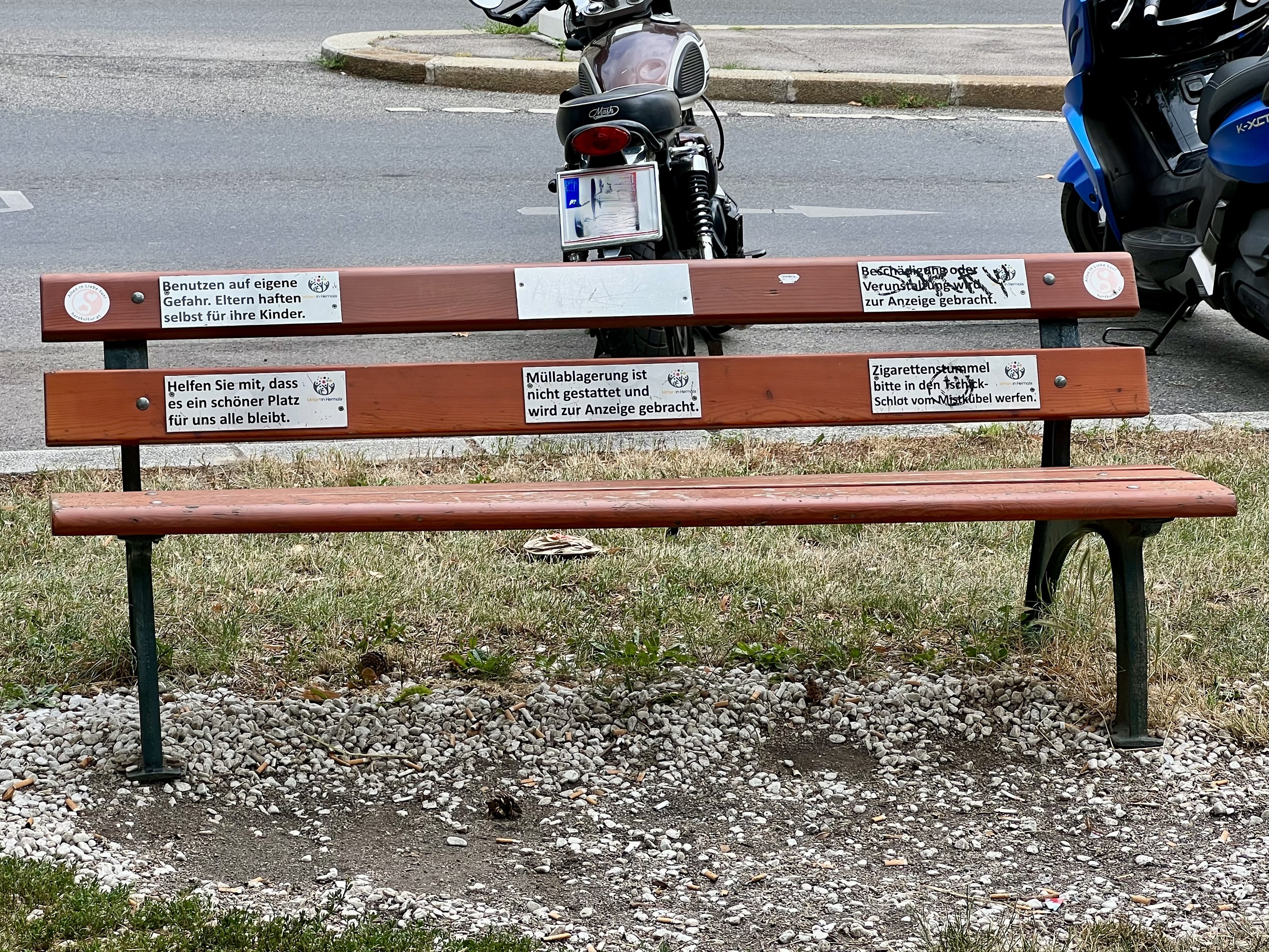 Eine hölzerne Parkbank mit mehreren Schildern, die verschiedene Anweisungen und Warnungen geben. Hinter der Bank sind geparkte Motorräder zu sehen. Die Bank steht auf einer Rasenfläche mit Kies und Zigarettenstummeln auf dem Boden.