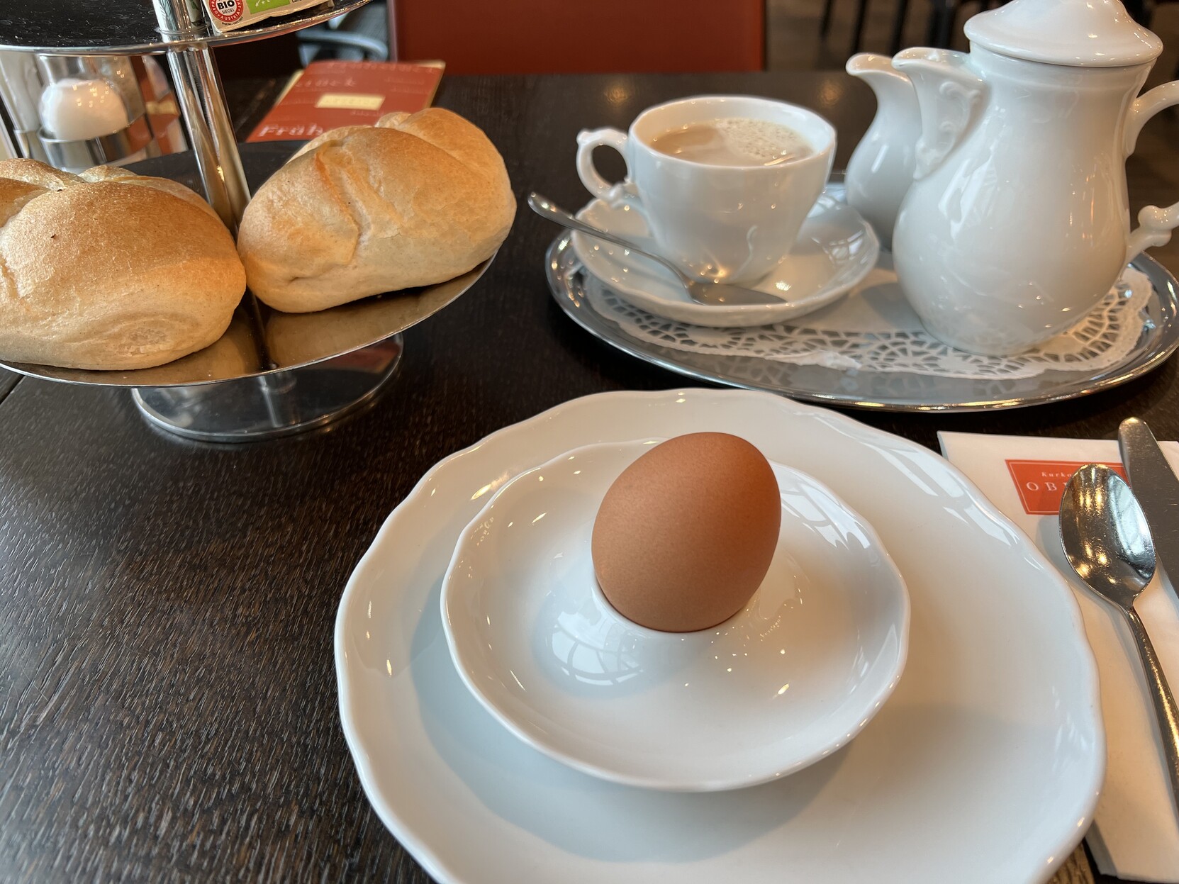 Tisch. Teller mit Ei. 2 Semmeln, Kännchen und Kaffeetasse. 