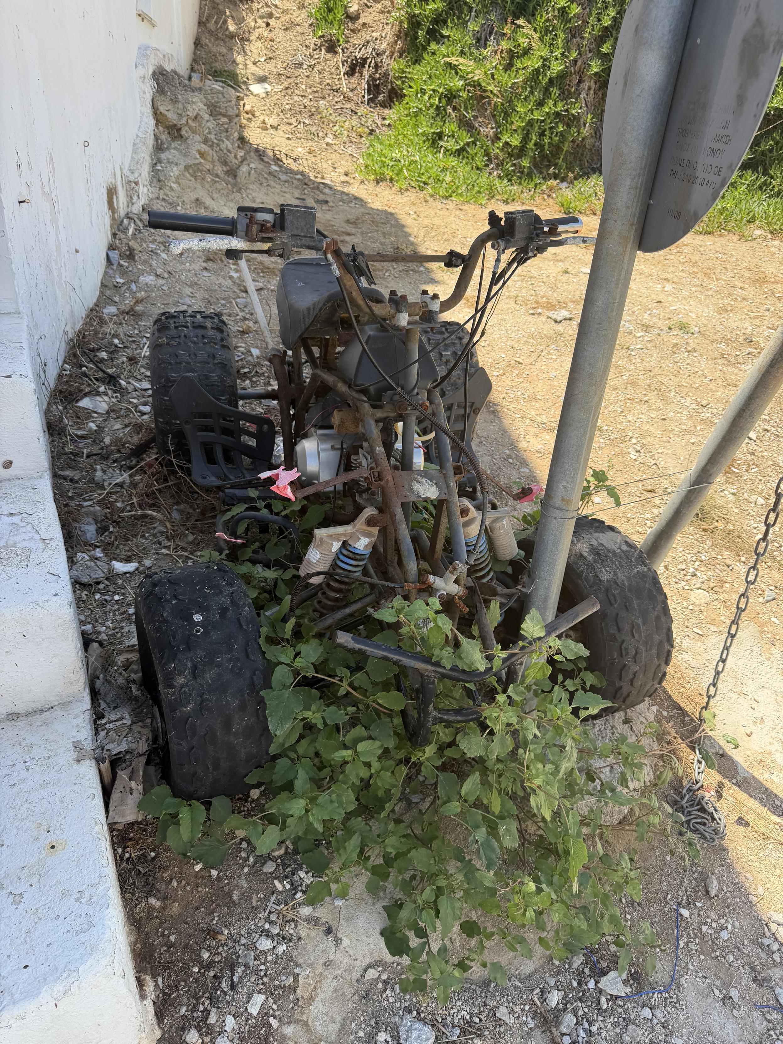 Ein alter, verrosteter Quad steht auf erdigen Gelände neben einem Verkehrszeichen und ist zum Teil mit Pflanzen überwuchert.