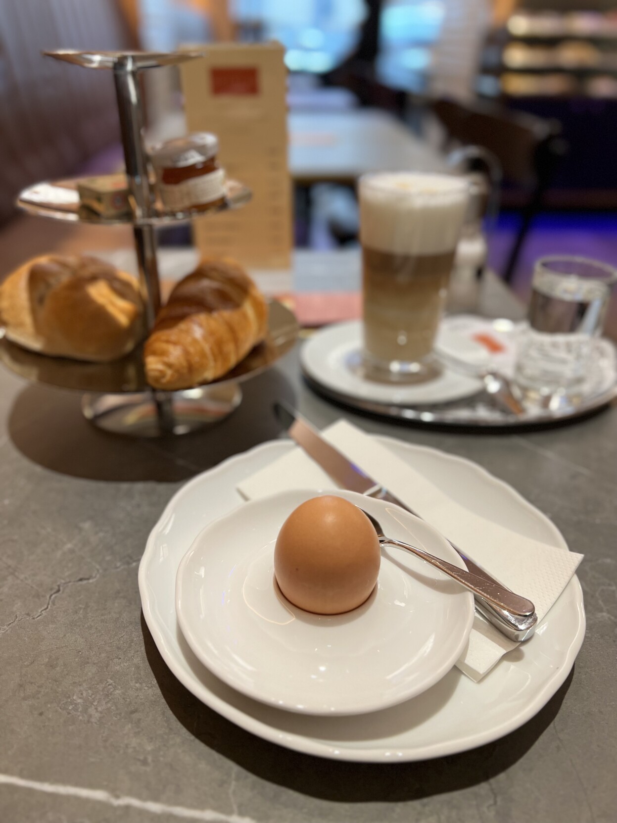 Teller mit Ei. Gestell mit Gebäck. Glas Café Latte. Alles auf einem Tisch. 