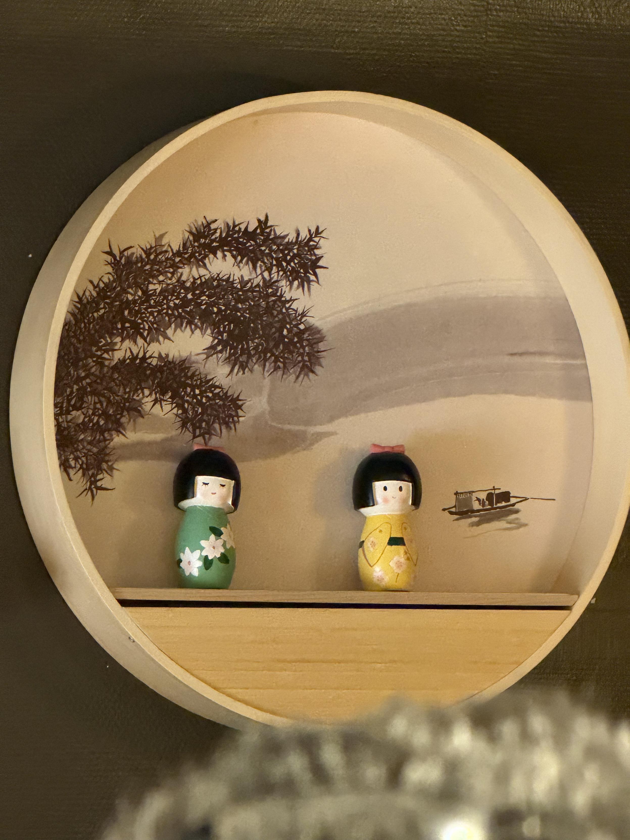 Rundes Wandregal mit zwei kleinen japanischen Kokeshi-Puppen in Grün und Gelb vor einer bemalten Landschaft mit Baum und Boot.