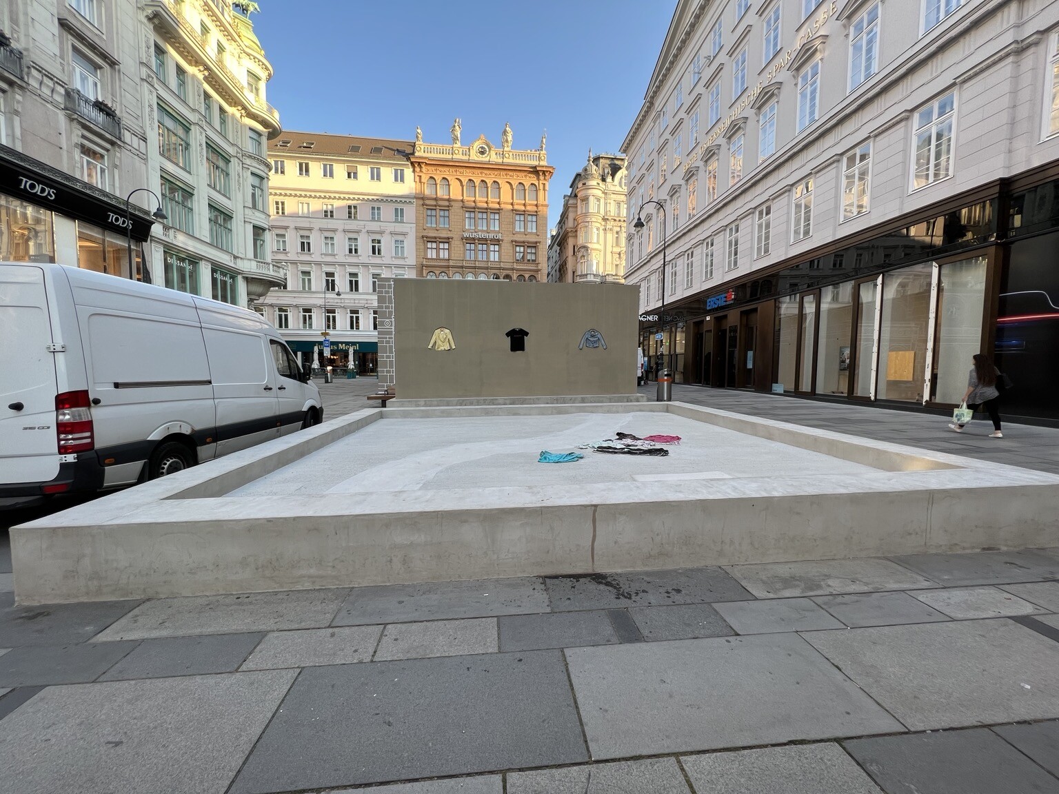 Große Betonplatte auf der ein paar Kleidungsstücke liegen. Dahinter Betonmauer in der drei Oberbekleidungen eingefasst sind.