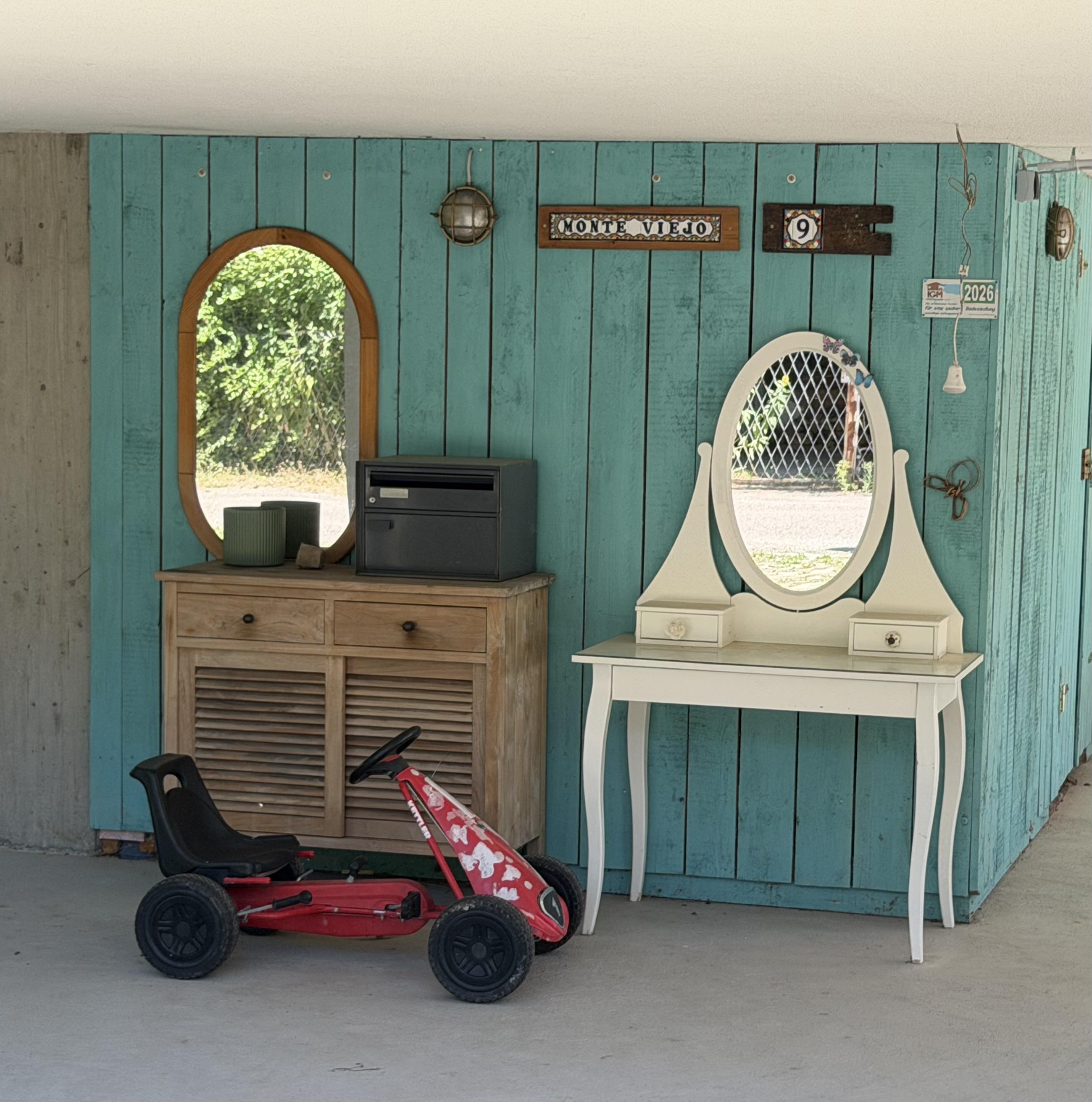 Ein rotes Kindertrettauto steht vor einer hellblauen Holzwand mit Spiegel und Dekorationen.Auf einer Kommode steht ein Postkasten. Daneben steht ein alter Schminktisch.