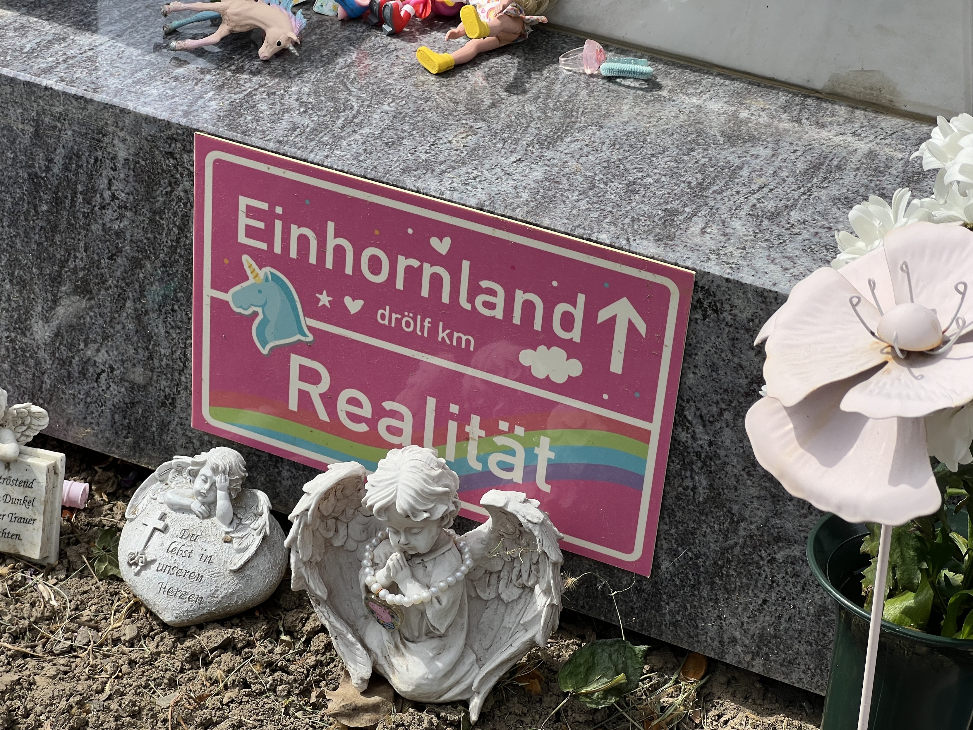 Ein Schild mit der Aufschrift „Einhornland“ und „drölf km Realität“ auf einem Sockel eines Grabsteins, umgeben von kleinen Engelsfiguren und Spielzeugen.