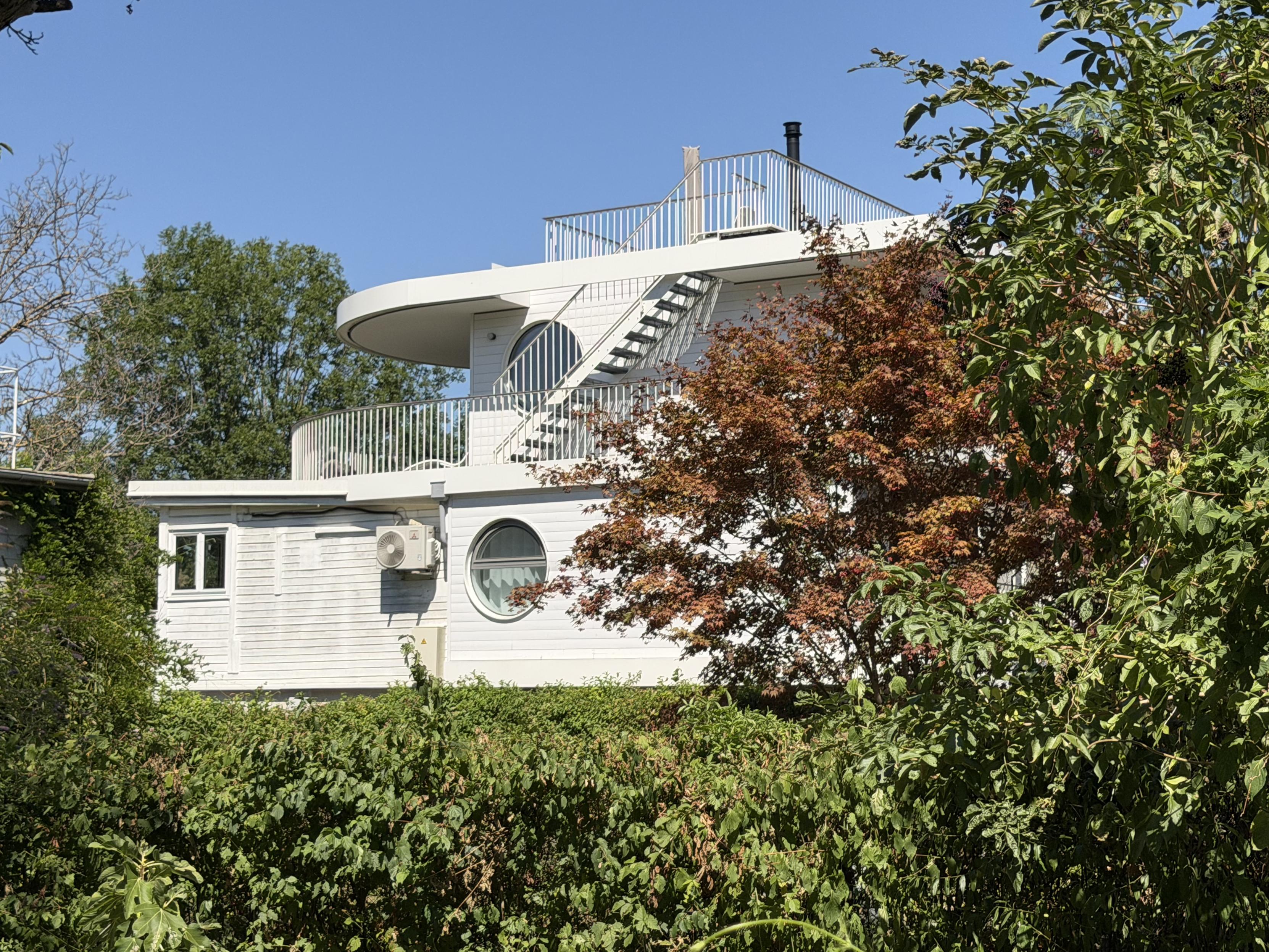 Modernes, weißes Haus mit runden Fenstern, Balkon und Außentreppe, umgeben von Bäumen und Sträuchern unter klarem blauen Himmel. Das Haus wirkt ein wenig wie der Aufbau eines Schiffes.