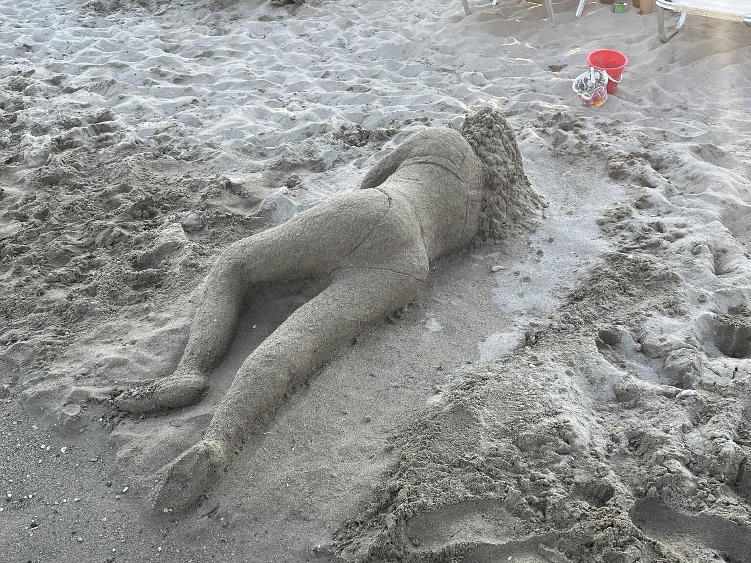 Liegende Frau in Bikini als Sandfigur - von hinten.