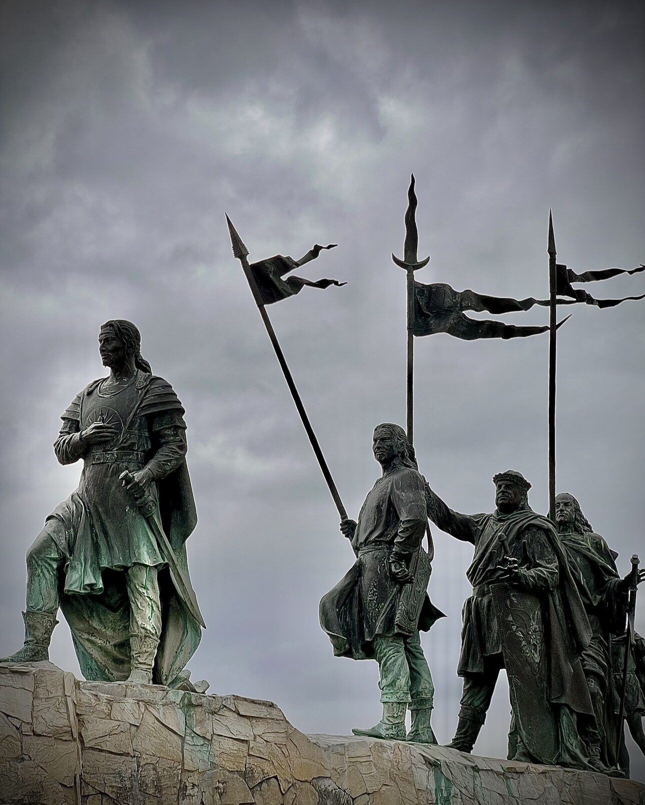 Vier Statuen von Männern in mittelalterlichen Gewändern hintereinander stehend. Der erste hält eine Hand an der Brust und die andere am Griff des Schwertes, dass in der Scheide steckt. Die anderen halten Lanzen hoch an denen kleine Fahnen wehen. 