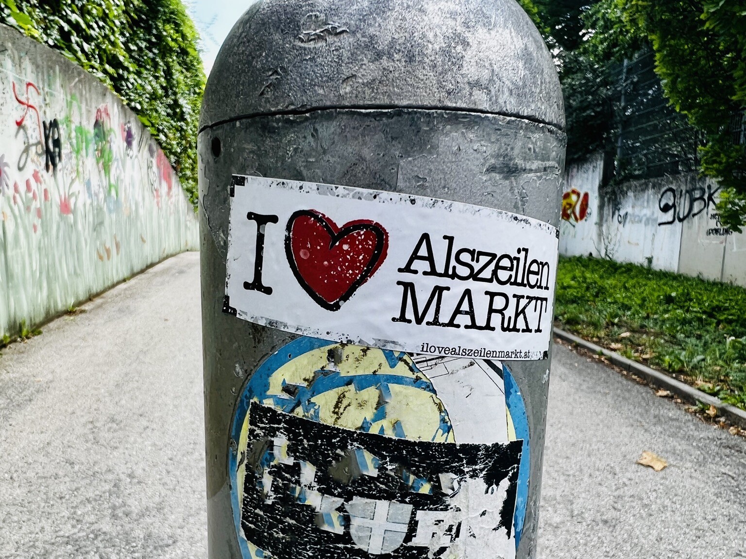 Gasse. Im Vordergrund Pfosten mit Aufkleber „I ♥️ Alszeilenmarkt