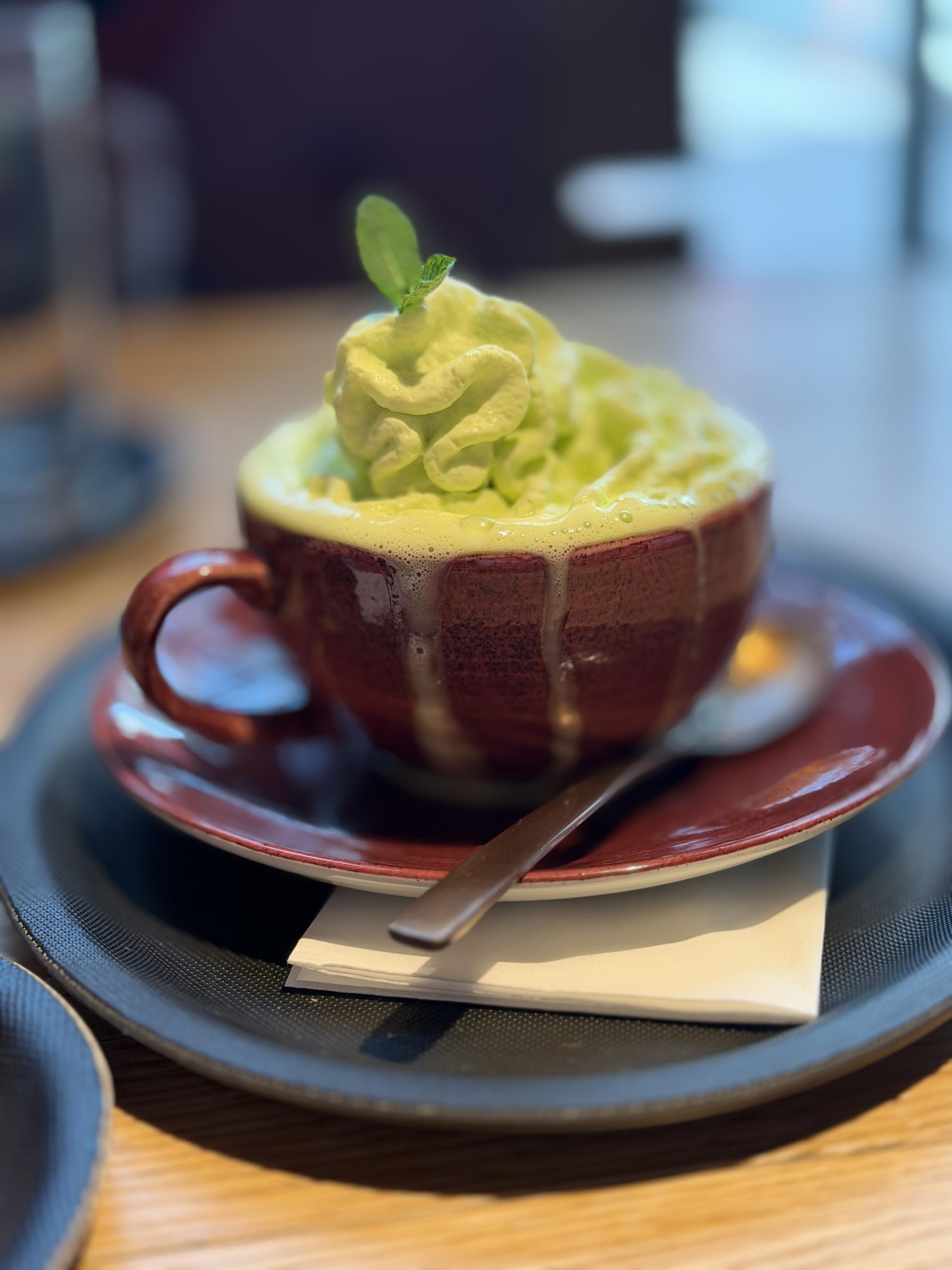 Eine braune Tasse mit cremigem Matcha-Getränk und hellgrünem Sahnetopping steht auf einer Untertasse mit Löffel auf einem Tablett im Café.