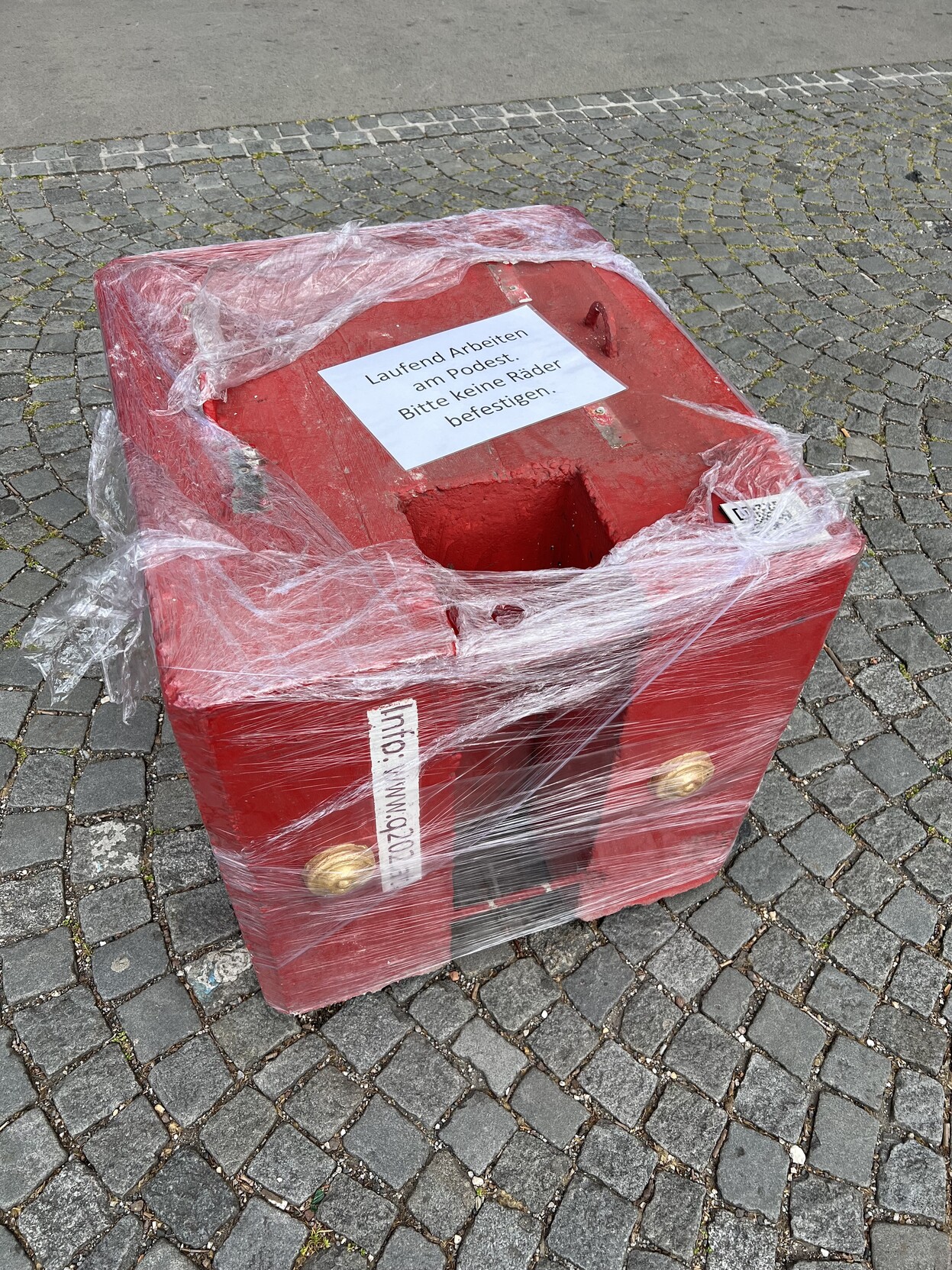 Roter Betonwürfel auf Gehsteig. Er ist in Plastik verpackt. Auf ihm ist ein Zettel aufgeklebt: „Laufende Arbeiten am Podest. Bitte keine Räder befestigen.“