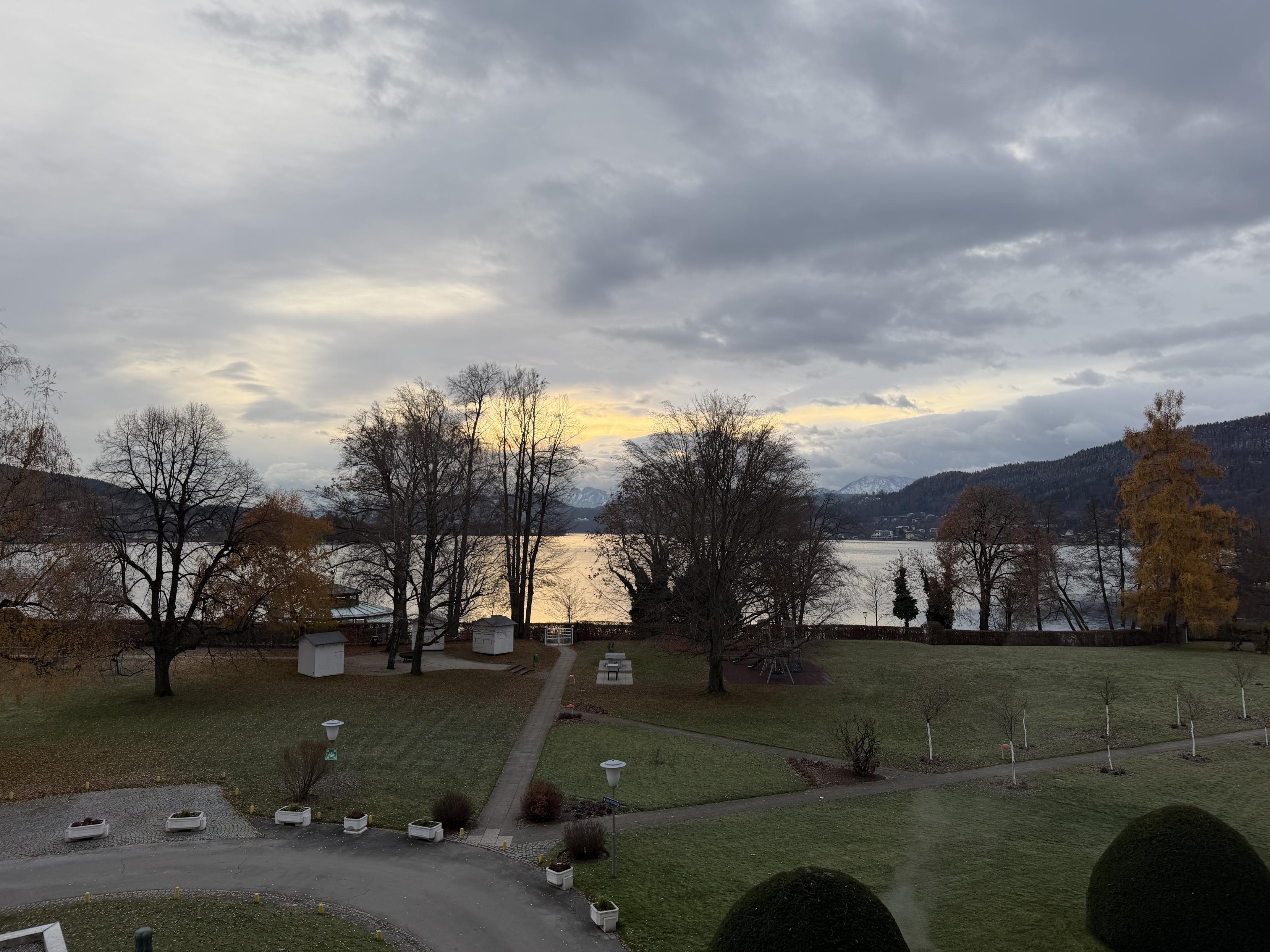 Weitläufiger Blick über eine herbstliche Parkanlage mit kahlen und teils goldgelben Bäumen, im Hintergrund ein ruhiger See und dahinter dunkle Hügel und schneebedeckte Berge unter einem bewölkten, grau-gelben Morgenhimmel.