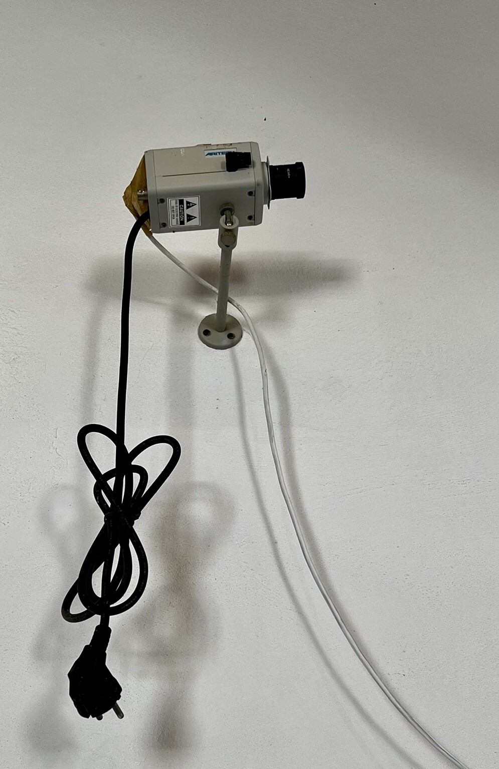 Überwachungskamera schaut Wand entlang schräg nach oben. Stromstecker hängt an Kabel in der Luft.