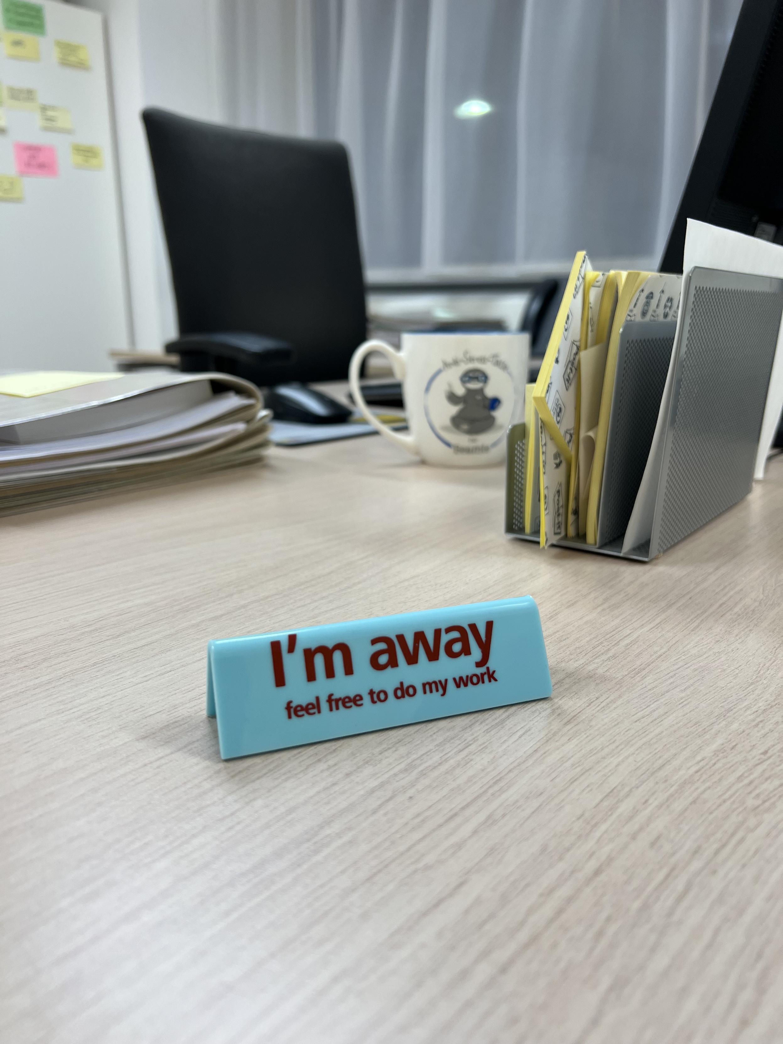 Ein Schreibtisch mit Büromaterialien, darunter Aktenstapel, eine Tasse und ein Monitor. Im Vordergrund steht ein Schild mit der Aufschrift „I'm away – feel free to do my work“. Im Hintergrund ist ein leerer Bürostuhl zu sehen.