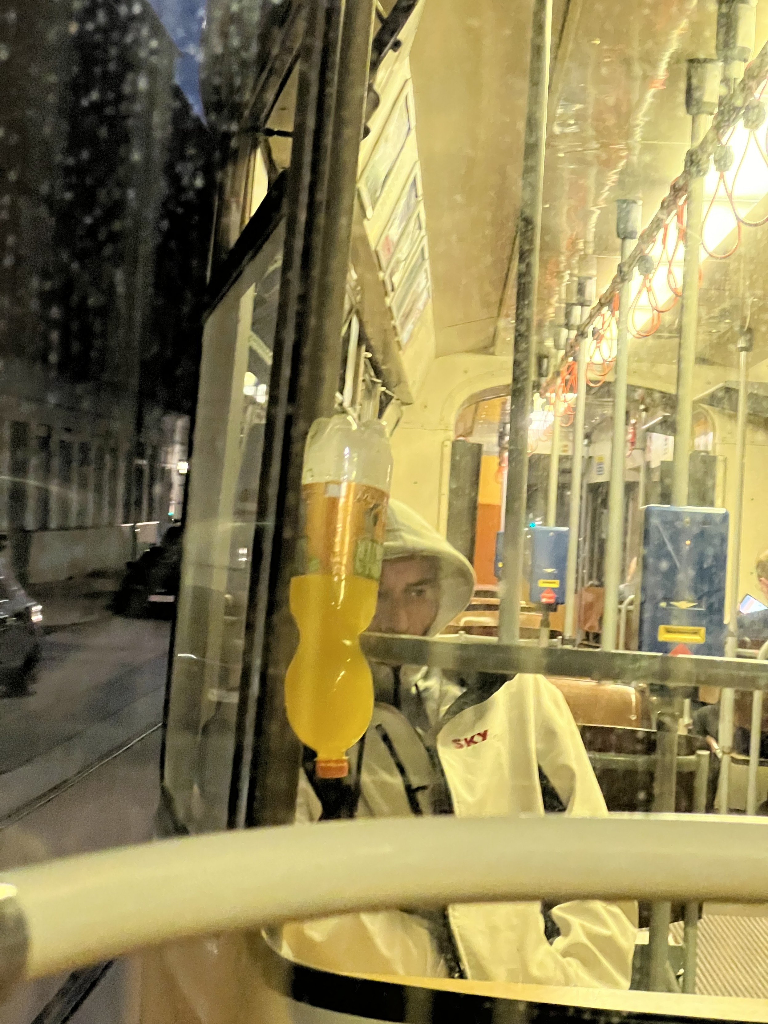 Blick in eine Straßenbahn aus dem hinteren Waggon heraus. Innenraum ist beleuchtet. Man sitzt mit Jacke und hochgezogener Kapuze am Wagenende. Dort ist in eine Haltestange eine große Limonadenflasche mit dem Verschluss nach unten eingeklemmt. Sie ist mit einer orangen Flüssigkeit gefüllt. 