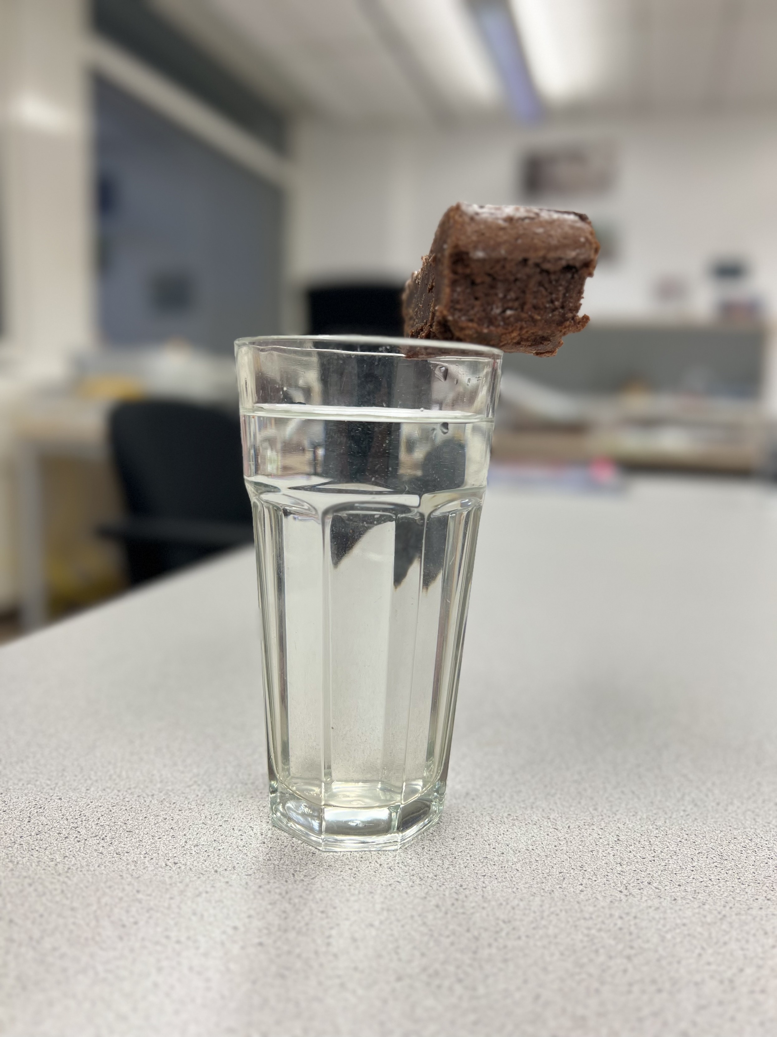 Ein Glas mit klarem Wasser steht auf einer Tischplatte, wobei der Fokus auf einem schwebenden Stück Schokoladenkuchen liegt, das unscharf im Vordergrund über dem Glas zu schweben scheint. Der Hintergrund ist unscharf, mit Elementen eines Klassenraums oder Büros.