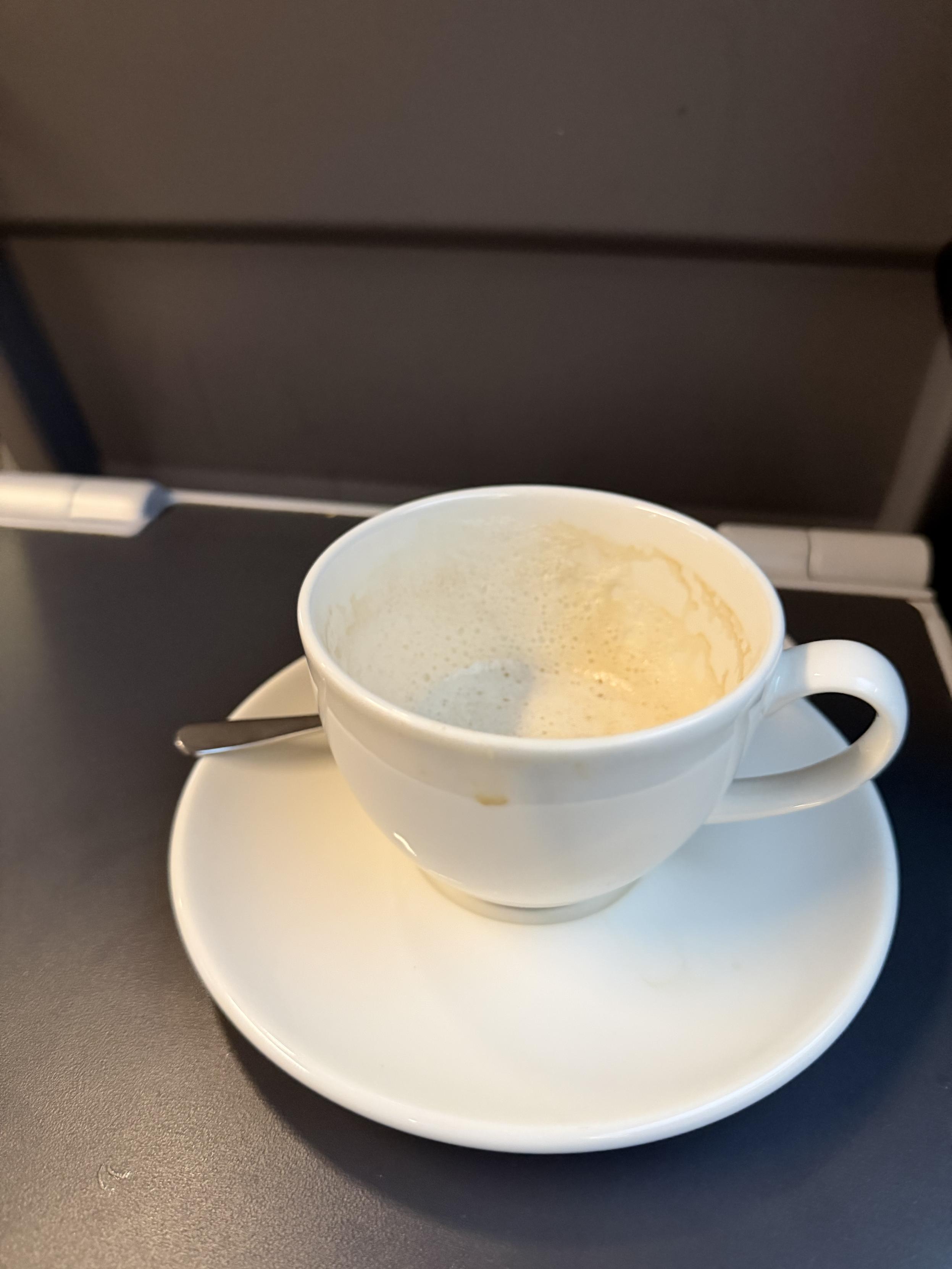 Weiße Kaffeetasse mit Milchschaumresten auf einer Untertasse und einem kleinen Löffel, stehend auf einem dunklen Klapptisch in einem Zug.