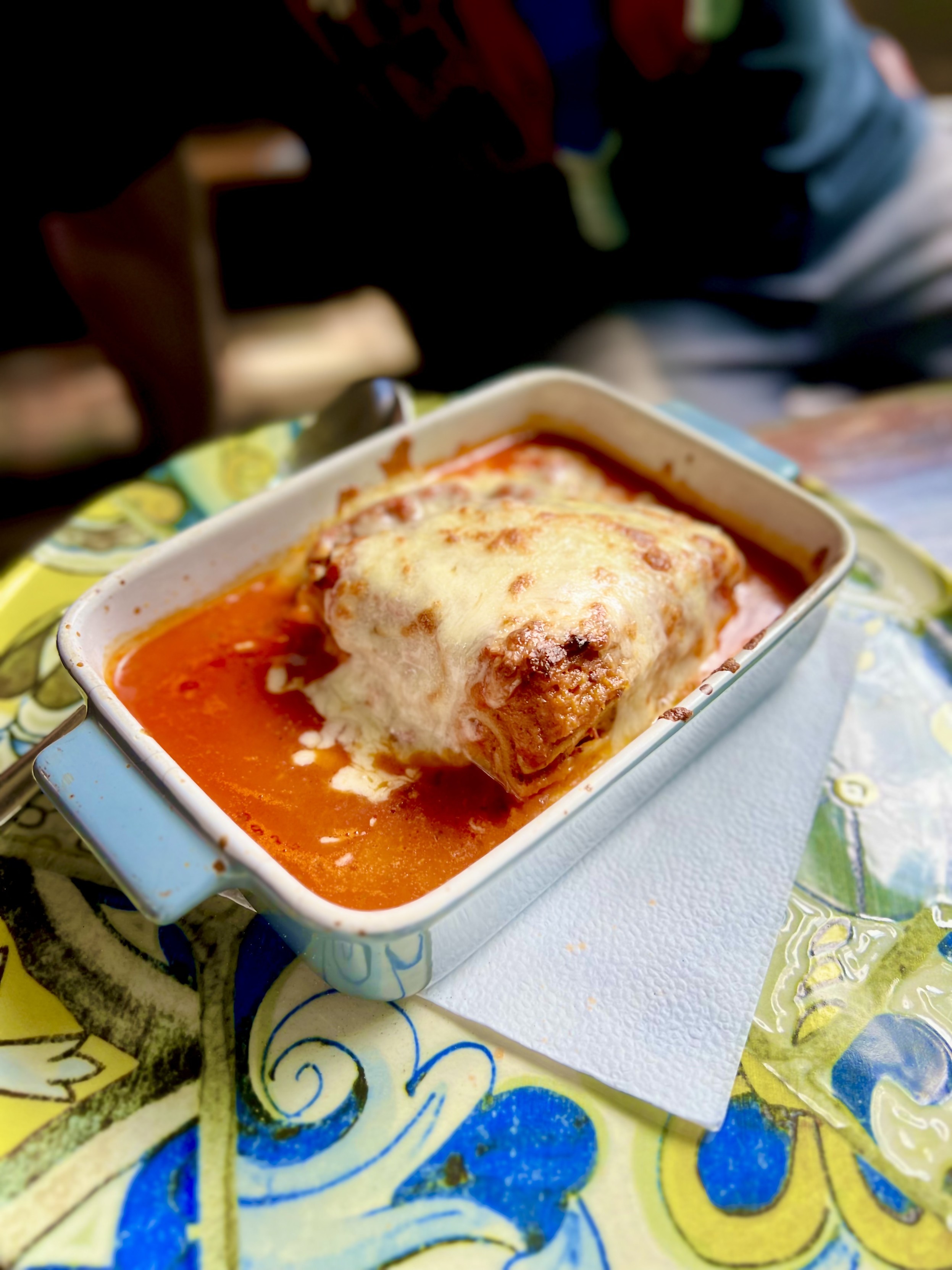 Eine Portion Lasagne, bedeckt mit geschmolzenem Käse, serviert in einer kleinen blauen Auflaufform auf einem bunt gemusterten Tisch.