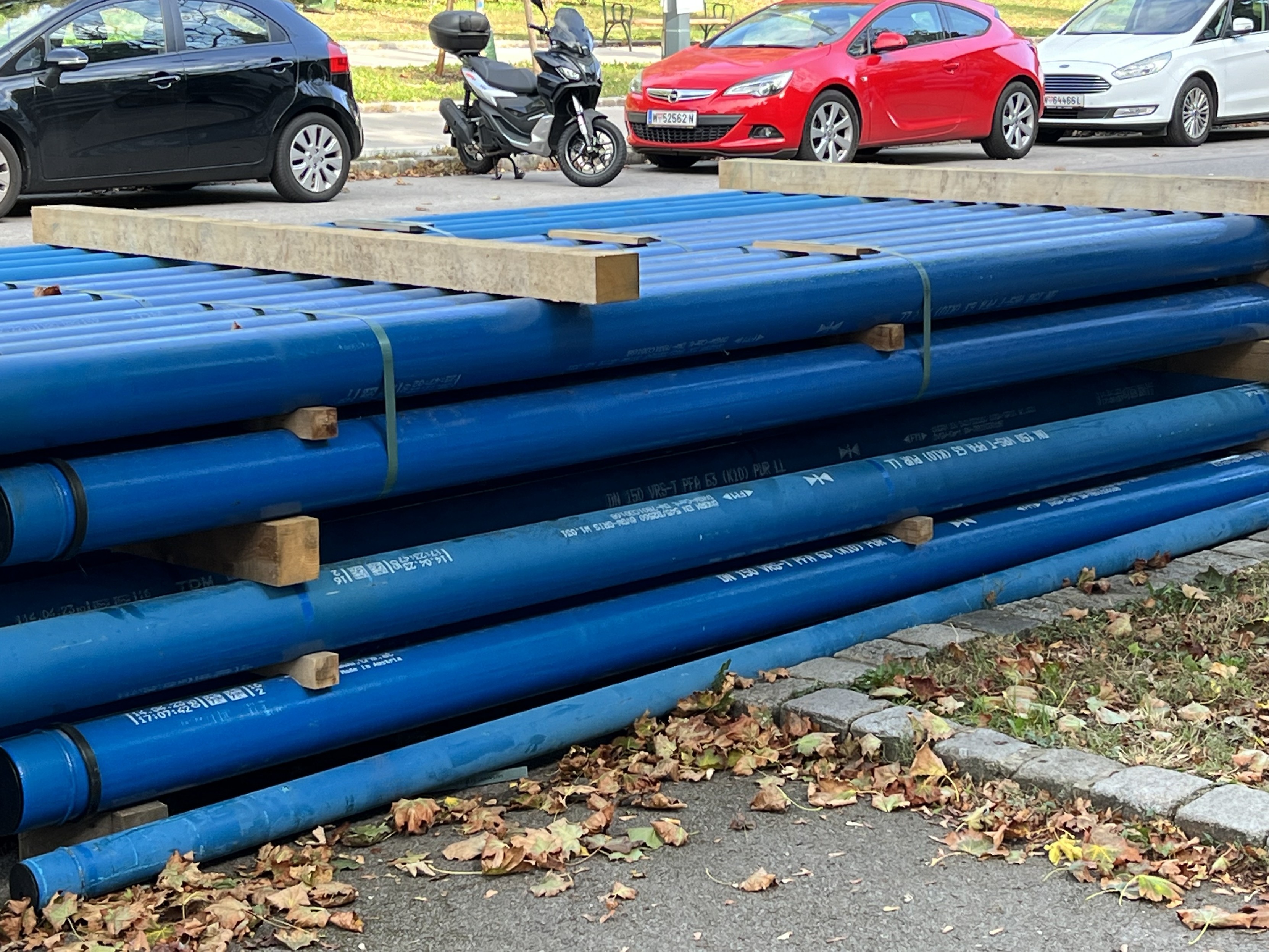 Stapel von blauen Rohren auf einer Baustelle mit Autos im Hintergrund.