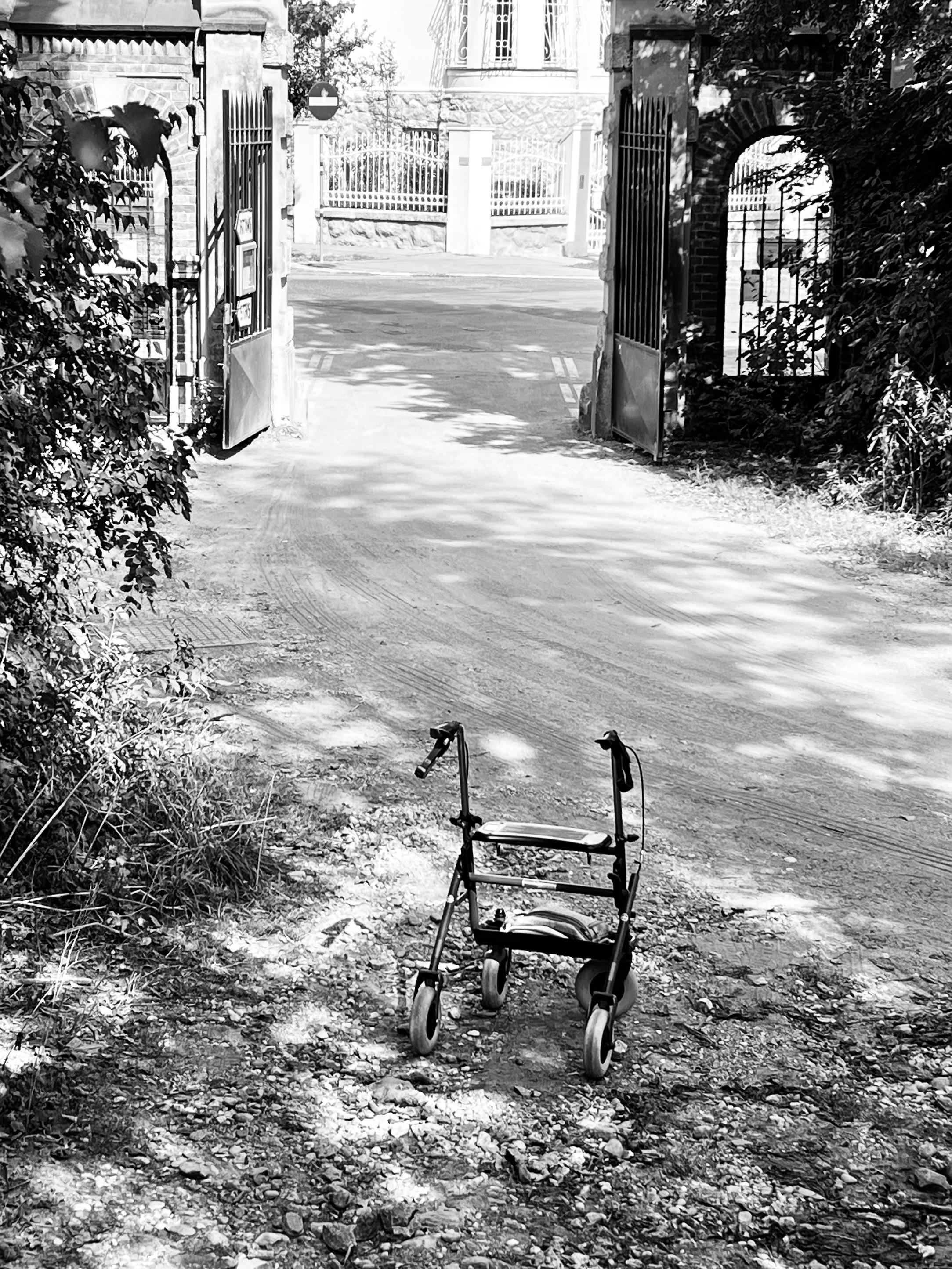 Ein verlassener Rollator steht auf einem unbefestigten Weg vor einem offenen Tor. Dahinter ist eine Einfahrt zu einem großen Gebäude mit dekorativem Zaun sichtbar. Die Szene ist in Schwarz-Weiß gehalten.