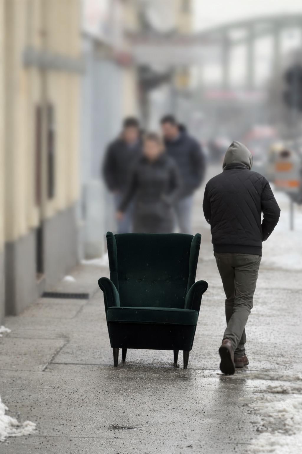 Ein grüner Ohrensessel steht auf einem nassen, verschneiten Gehsteig. Im Vordergrund geht eine Person mit Kapuze daran vorbei; im unscharfen Hintergrund sind drei weitere Personen zu sehen.