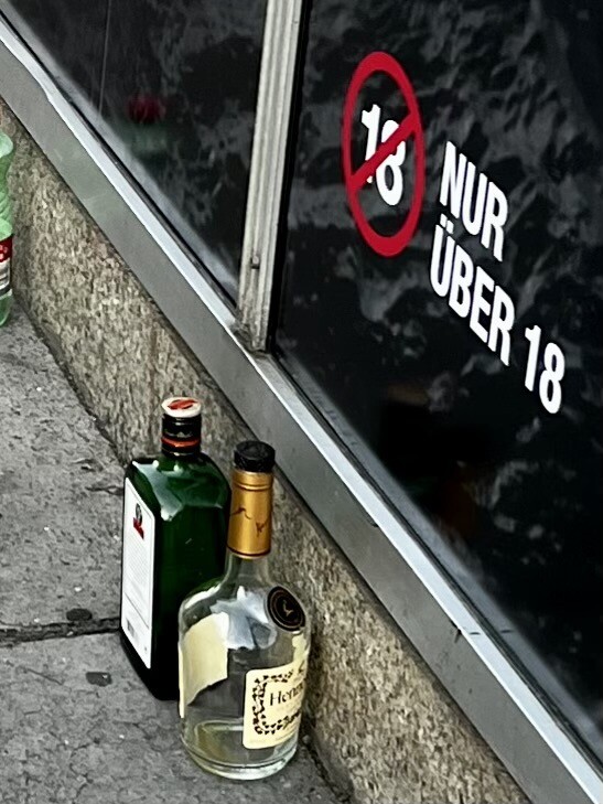 Zwei Flaschen mit hochprozentigen Alkohol am Gehsteig. Dahinter Schaufenster mit Schriftzug „Nur über 18“