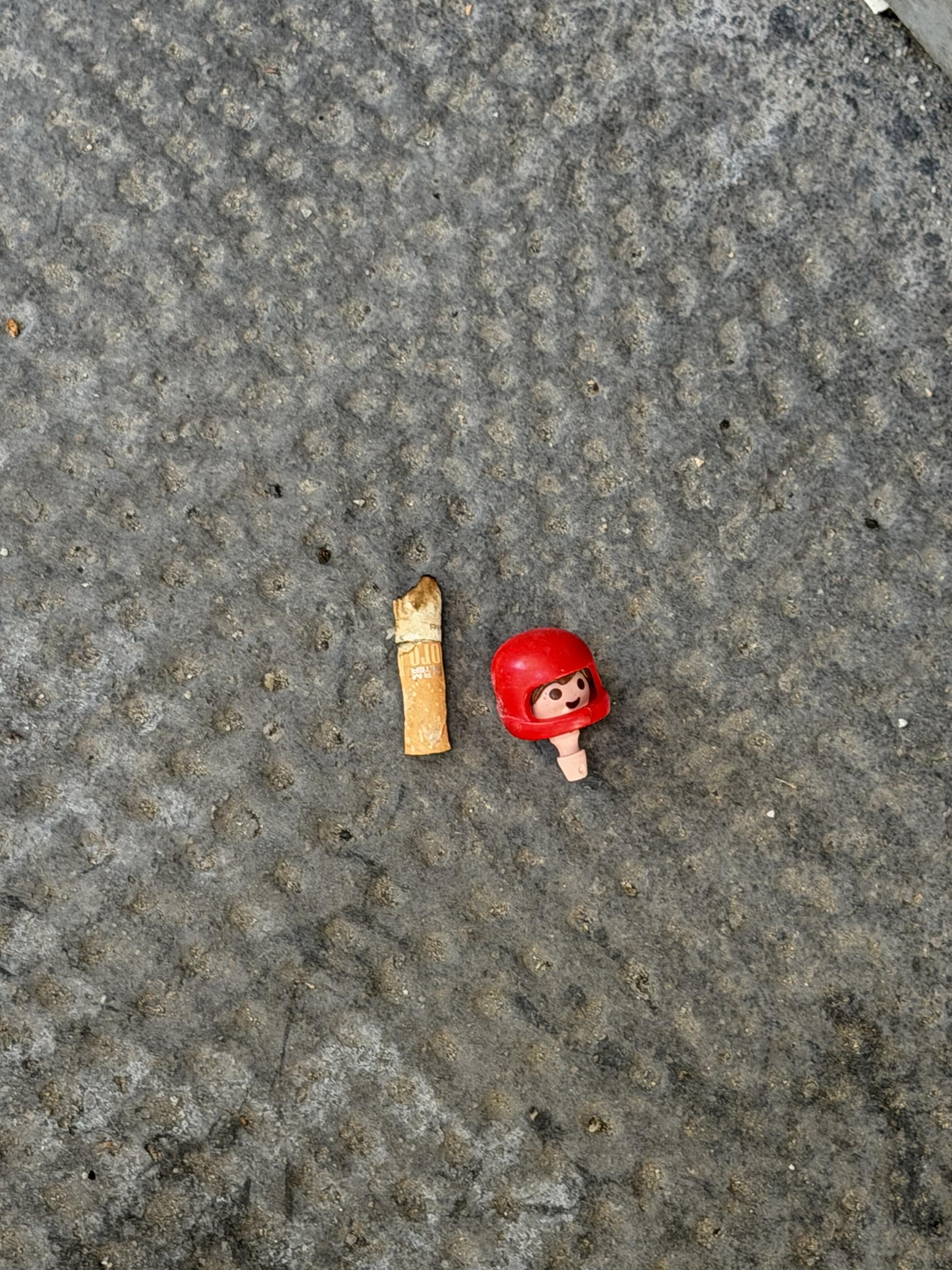 Auf einer grauen, rauen Asphaltoberfläche liegen nebeneinander ein weggeworfener Zigarettenstummel und der Kopf einer kleinen Spielfigur mit rotem Helm und lächelndem Gesicht.