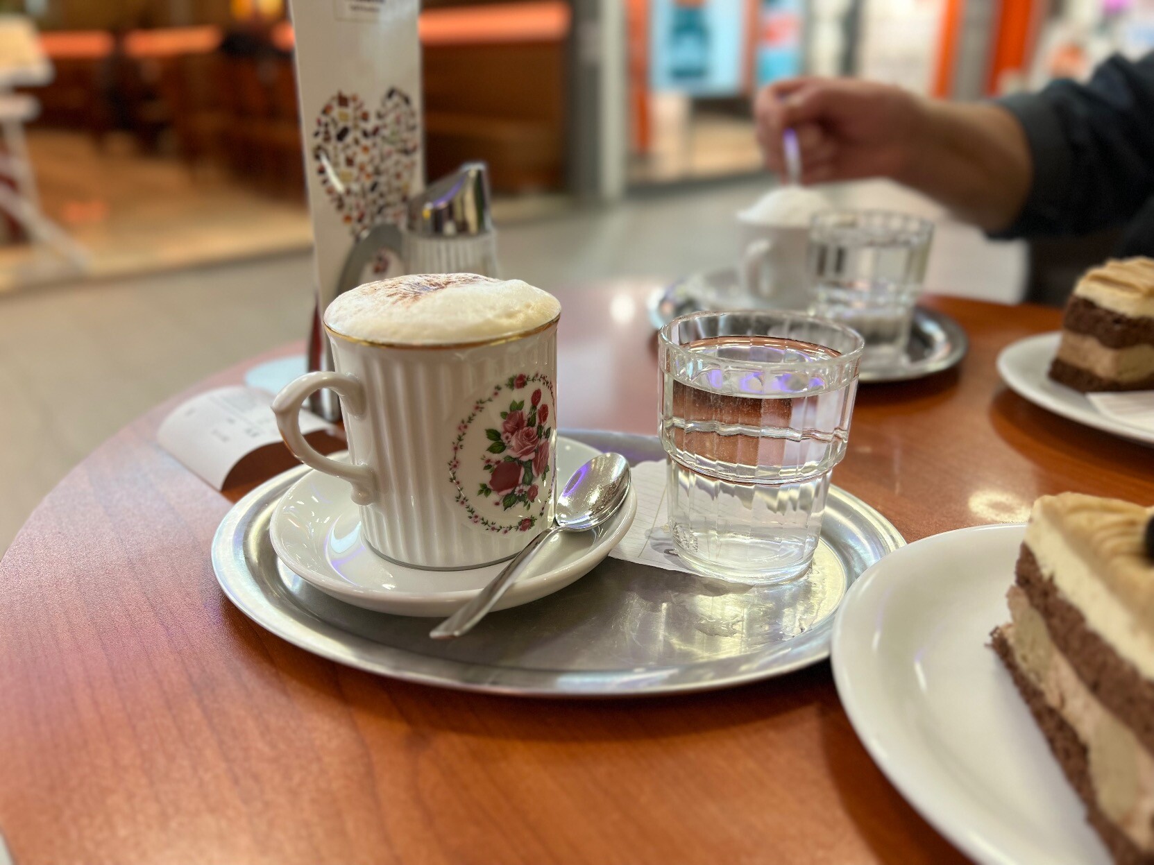 Tisch mit Tasse auf der Kaffeehäferl und Wasserglas steht. Daneben  ein Teil eines Tortenstücks zu sehen. Im Hintergrund Hand, die einem Kaffeehäferl mit Löffel umrührt.