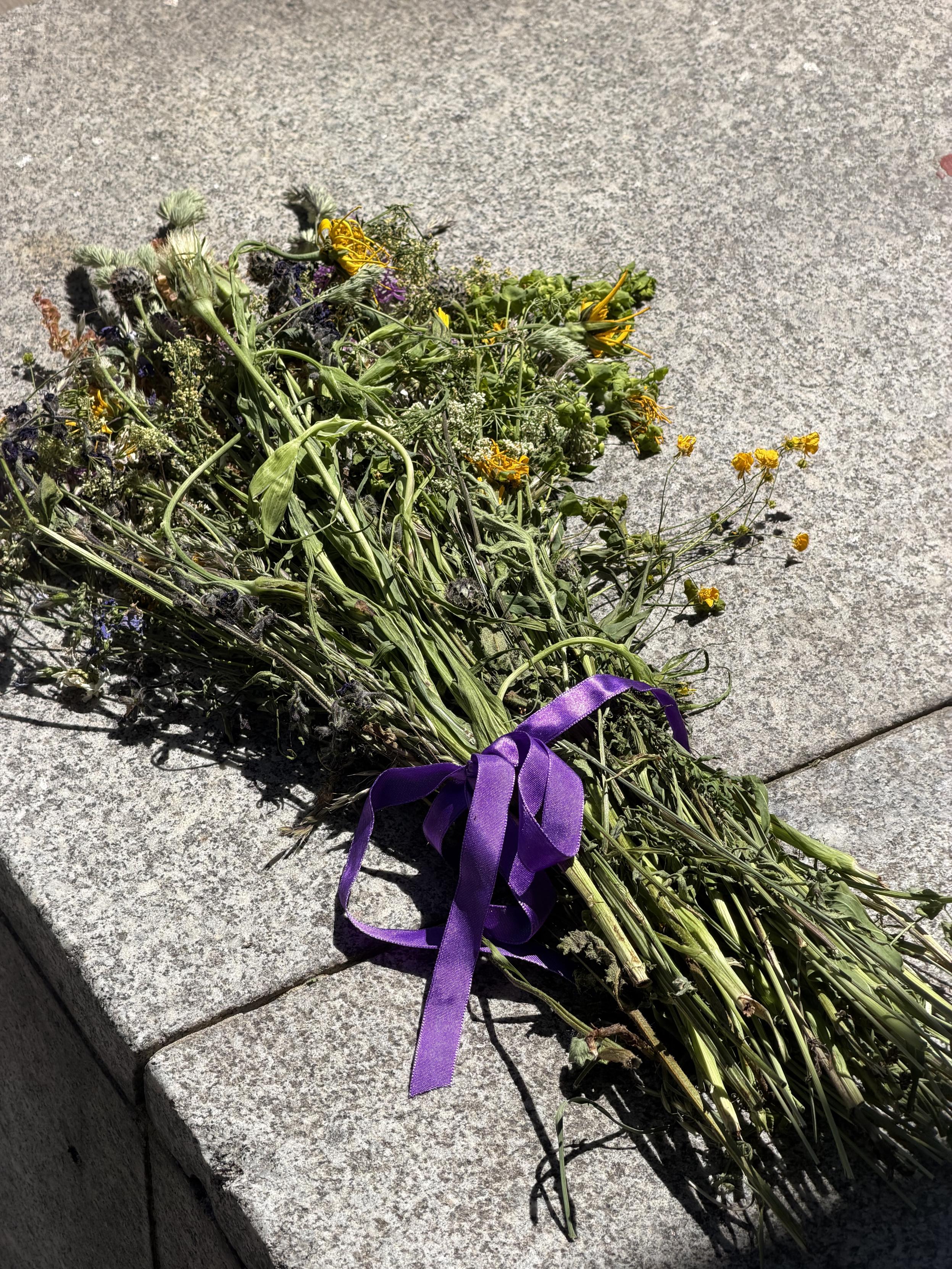 Ein verwelkter Wildblumenstrauß mit gelben, violetten und grünen Pflanzen liegt auf einer hellgrauen Steinfläche. Die Stiele sind mit einem violetten Band zusammengebunden und werfen in der Sonne deutliche Schatten.