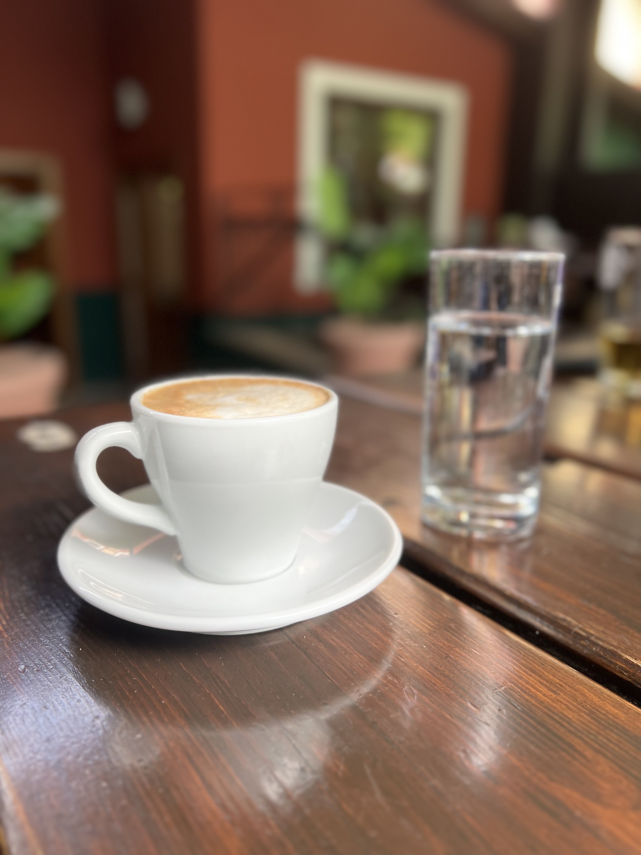 Eine Tasse Cappuccino auf einem Holztisch neben einem Glas Wasser in einem Lokal mit unscharfem Hintergrund.
