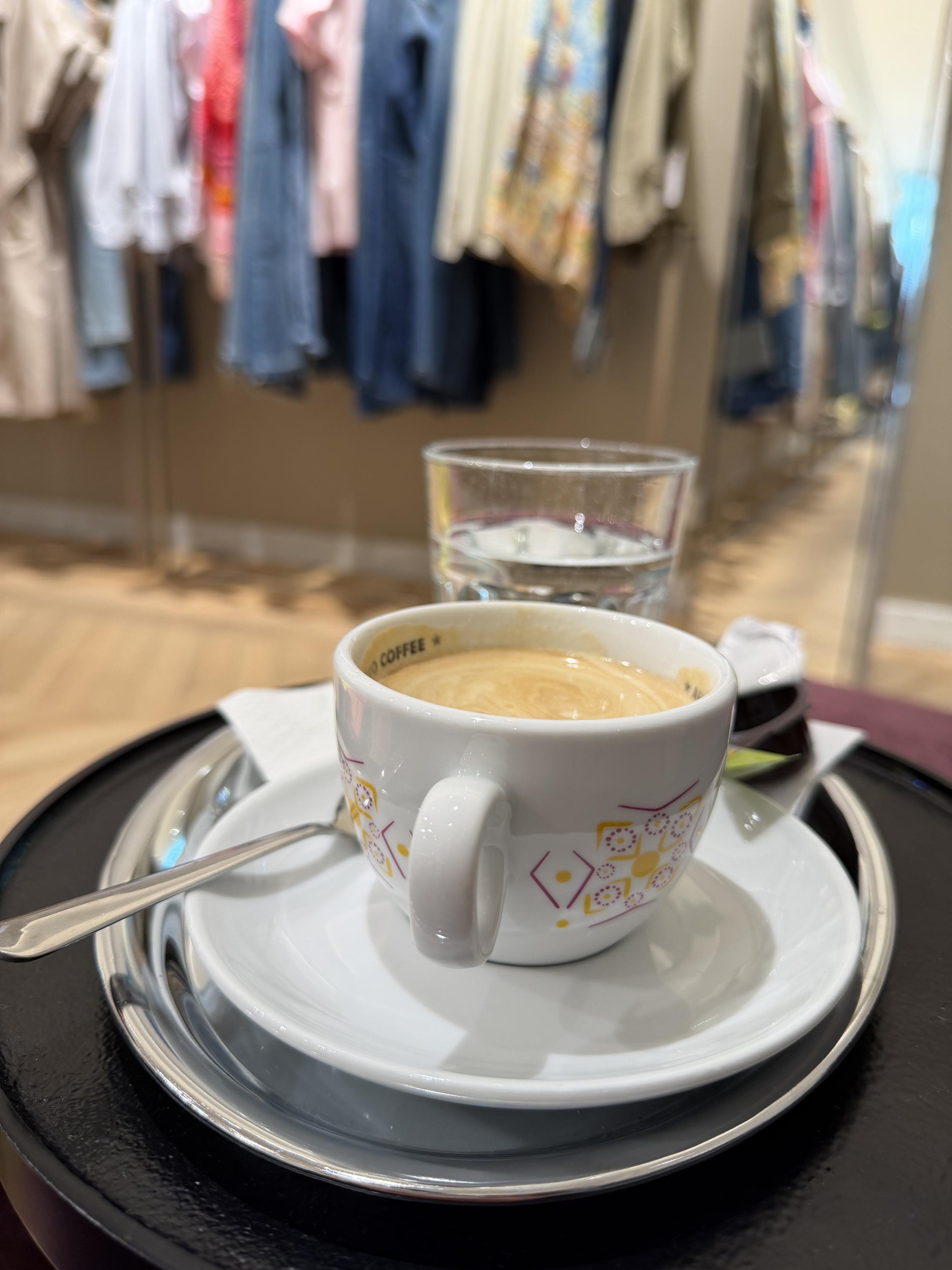 Kaffeehäferl mit Wasserglas auf kleinem Tischchen. Dahinter sieht man Kleidungsstücke an einer Stange aufgehängt.