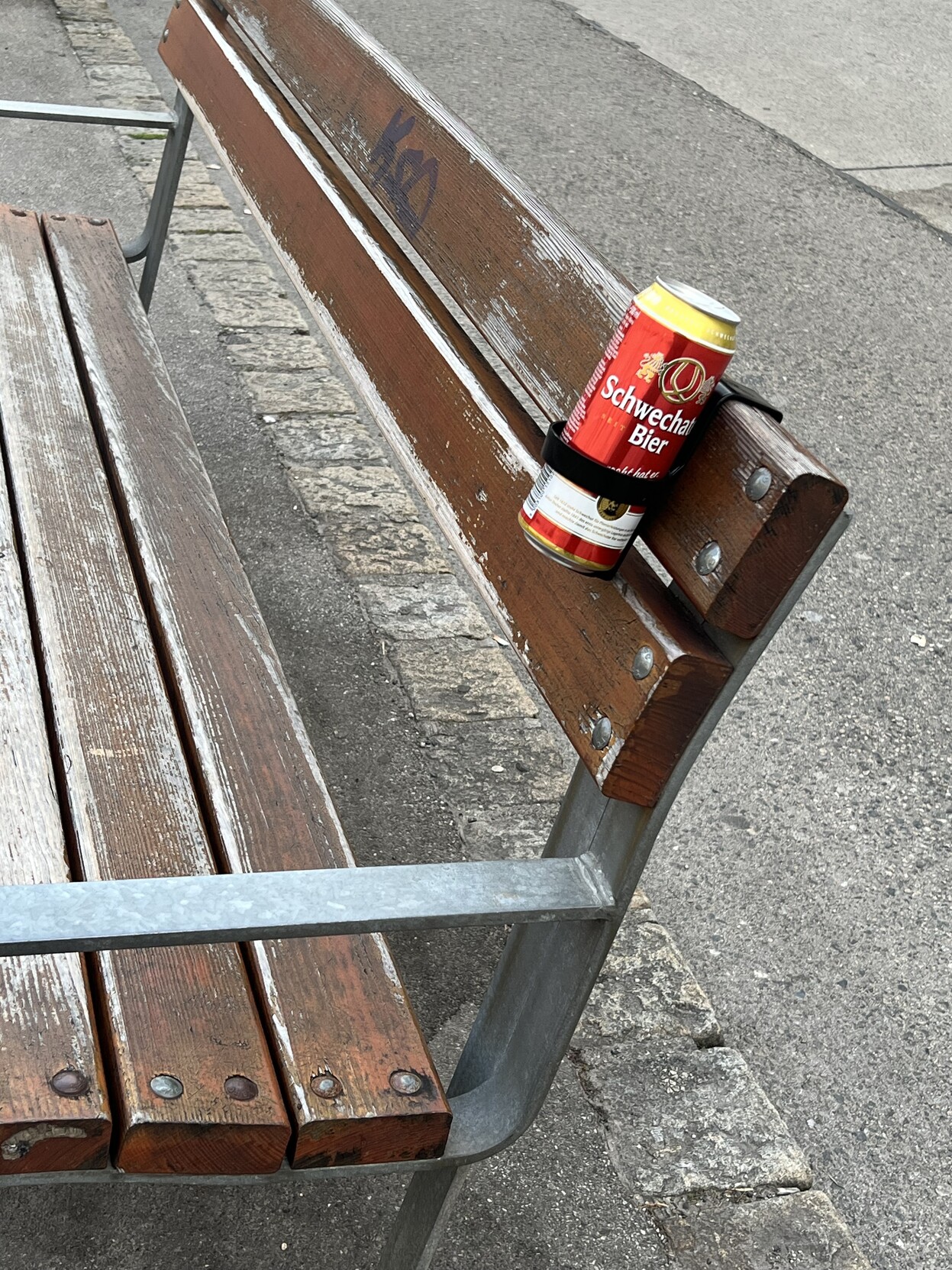 Sitzbank im öffentlichen Raum. An der Lehne ist eine Halterung angeklemmt, in der eine geöffnete Bierdose steckt. 