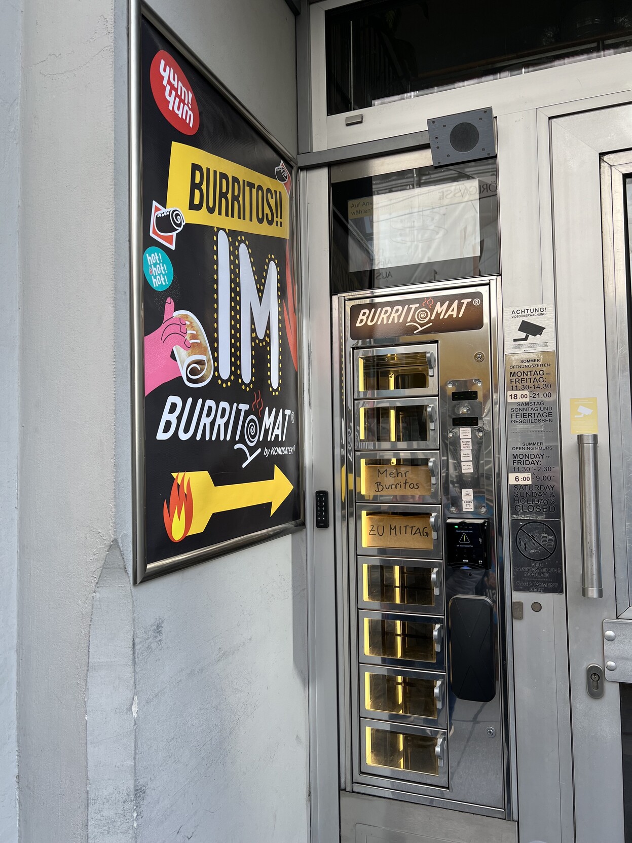 Werbeplakat an Hauswand 
: „Burritos im Burritomat“. Daneben ein Warenautomat „Burritomat“ mit Zettel „Mehr Burritos zu Mittag“