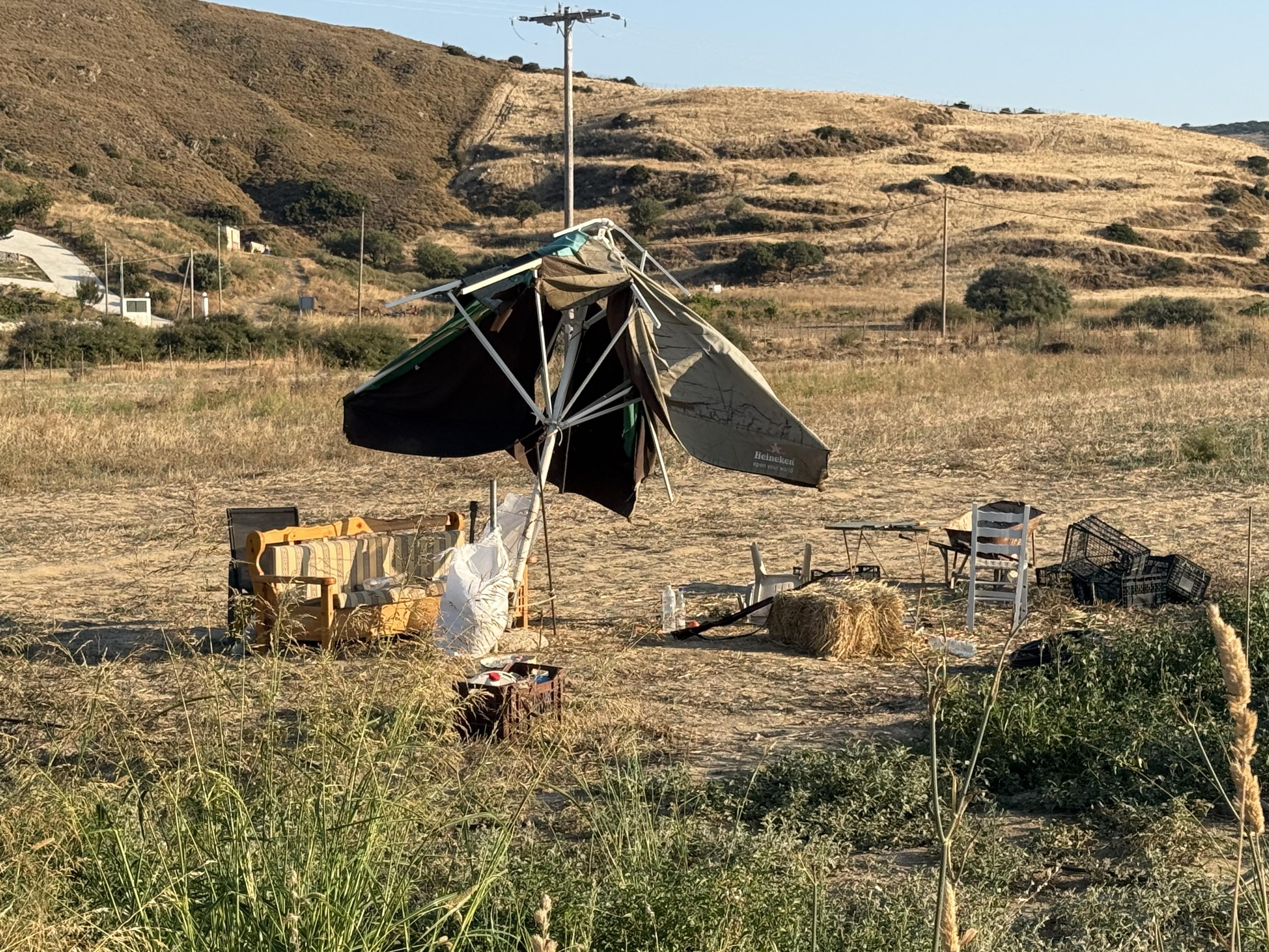 Abgeerntetes Feld. Kaputter aufgestellter Sonnenschirm. Altes Sofa. Heuballen. Unterschiedliche Stühle und Boxen.