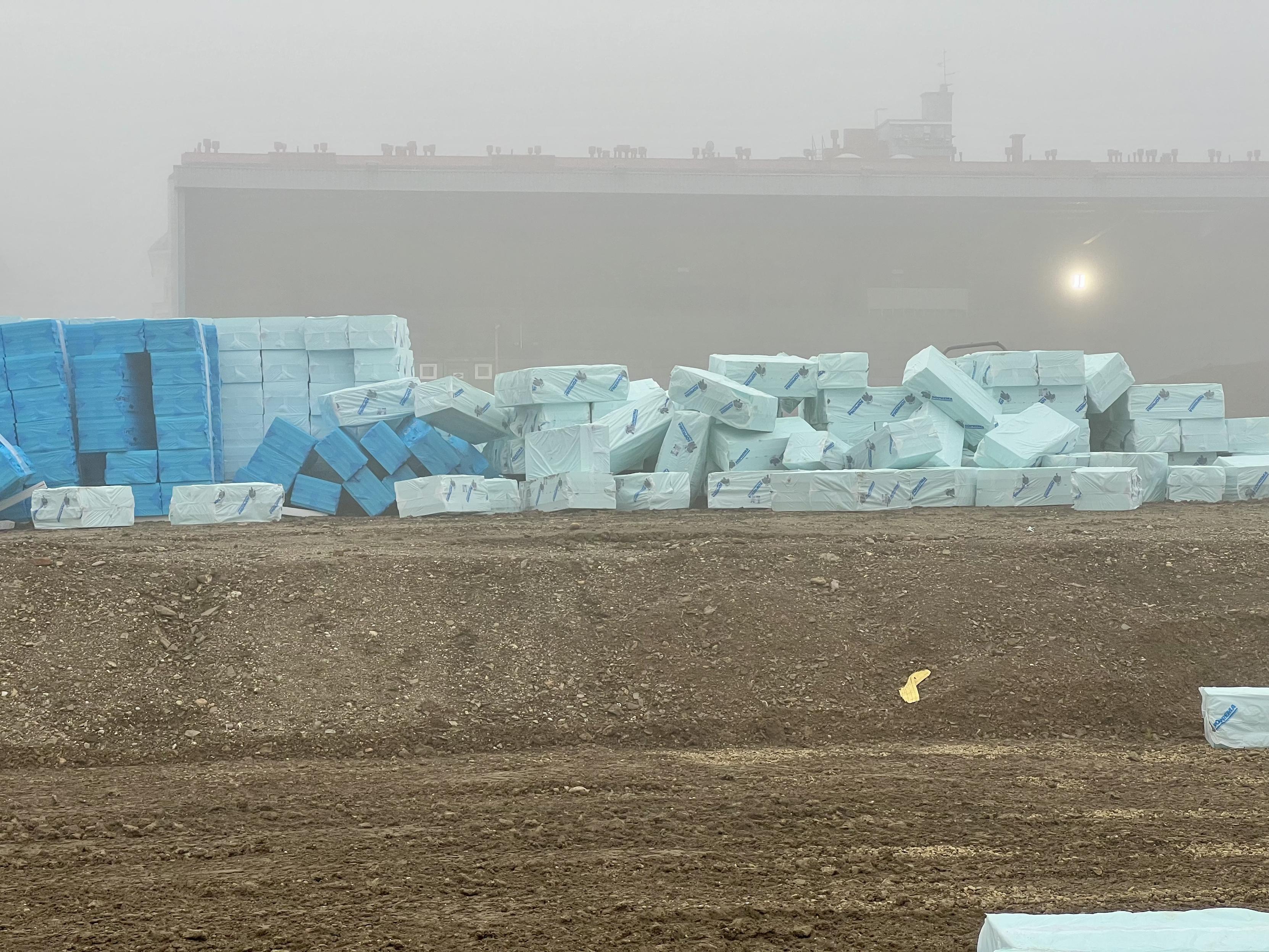 Ein Stapel von hellblauen, in Plastik verpackten Baumaterialien (vermutlich Dämmplatten) liegt auf einer Baustelle. Im Hintergrund ist ein großes, nebelverhangenes Gebäude zu erkennen. Der Boden im Vordergrund ist unbefestigt und erdig.