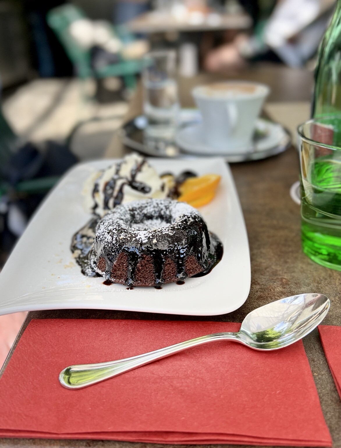 Im Vordergrund Löffel auf Serviette liegend. Dahinter kleiner Kuchen mit Schokolade und Staubzucker überzogen. Hinten am Teller Schlagobers und eine Orangenscheibe.