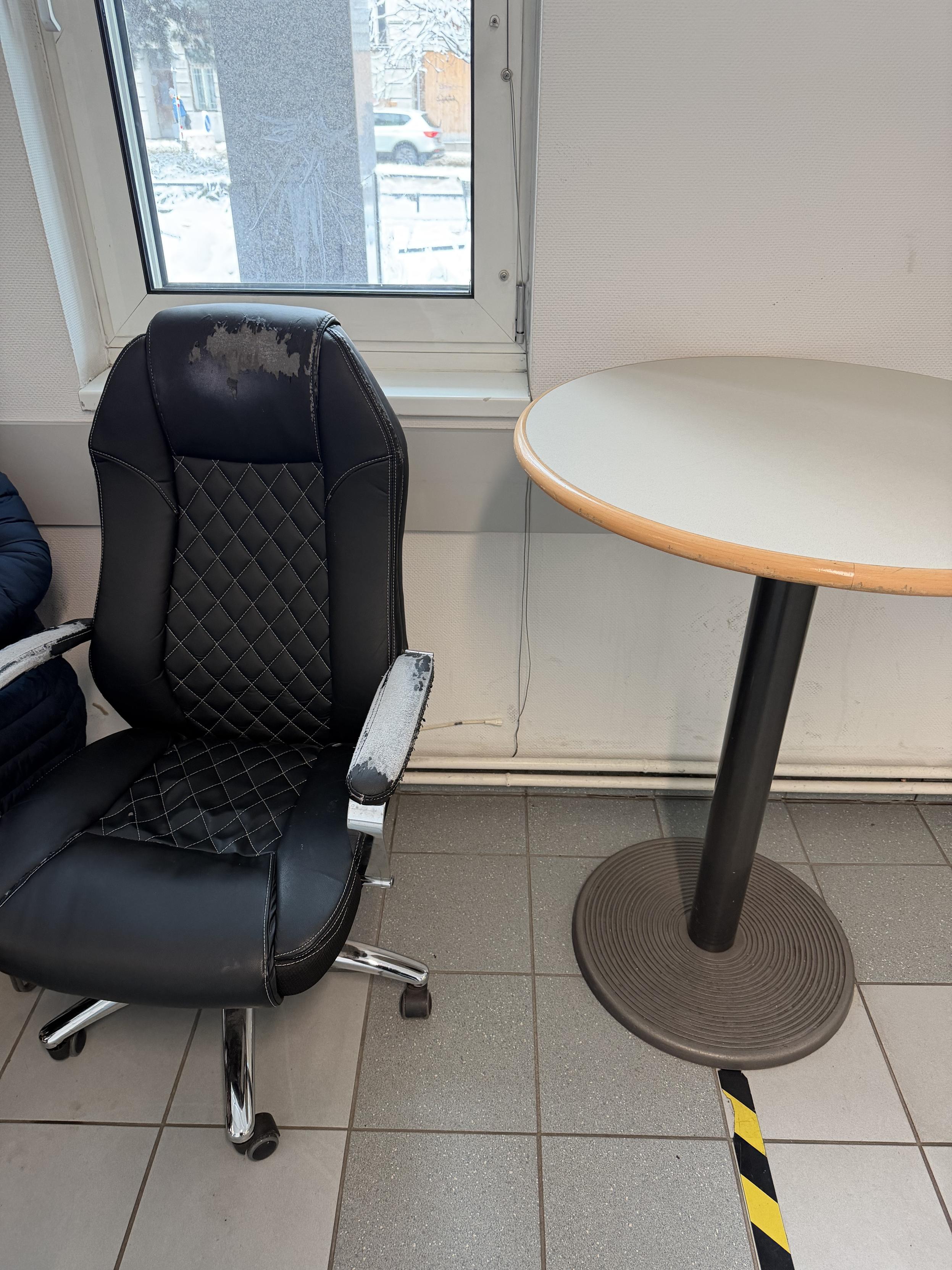 Schwarzer, stark abgenutzter Bürostuhl mit Rollen neben einem runden Stehtisch an einem Fenster; draußen ist eine verschneite Straße zu sehen, auf dem Fliesenboden verläuft ein gelb-schwarzer Markierungsstreifen.