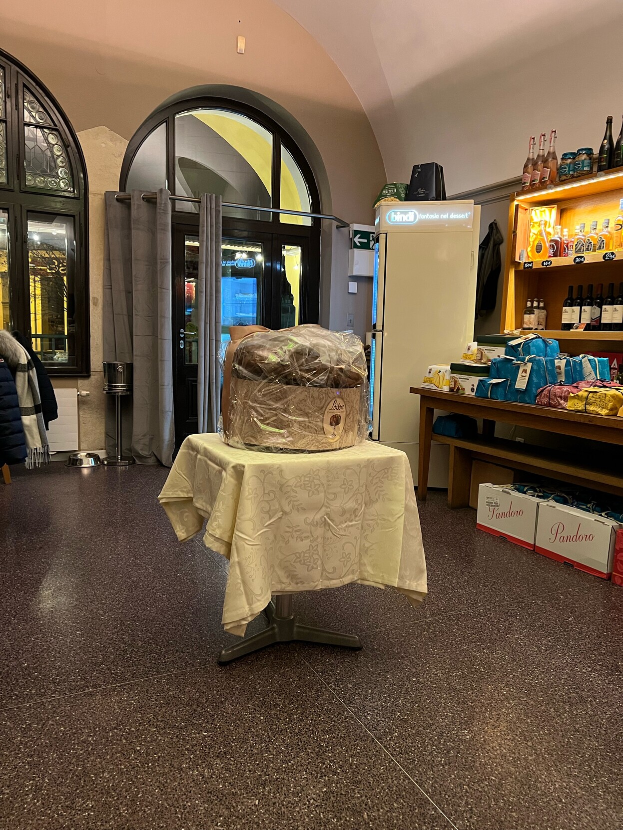 Großer Panettone steht auf einem kleinen Tischchen mitten in einem Lokal. Rechts Tisch und Regal mit kleineren Panettone und Flaschen.