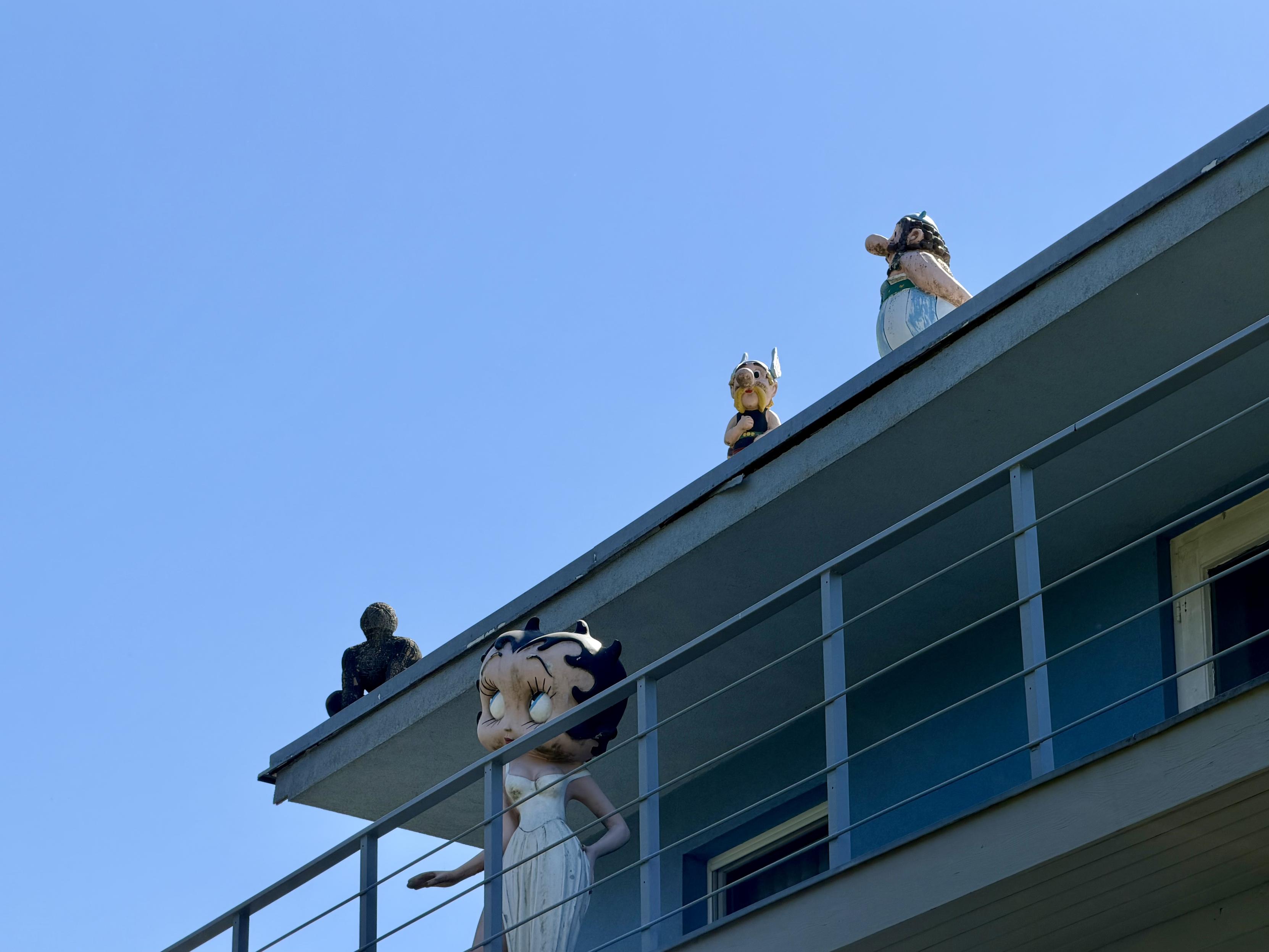 Figuren von Betty Boop, Buddha, Asterix und Obelix auf dem Balkon und Dach eines Gebäudes unter blauem Himmel.