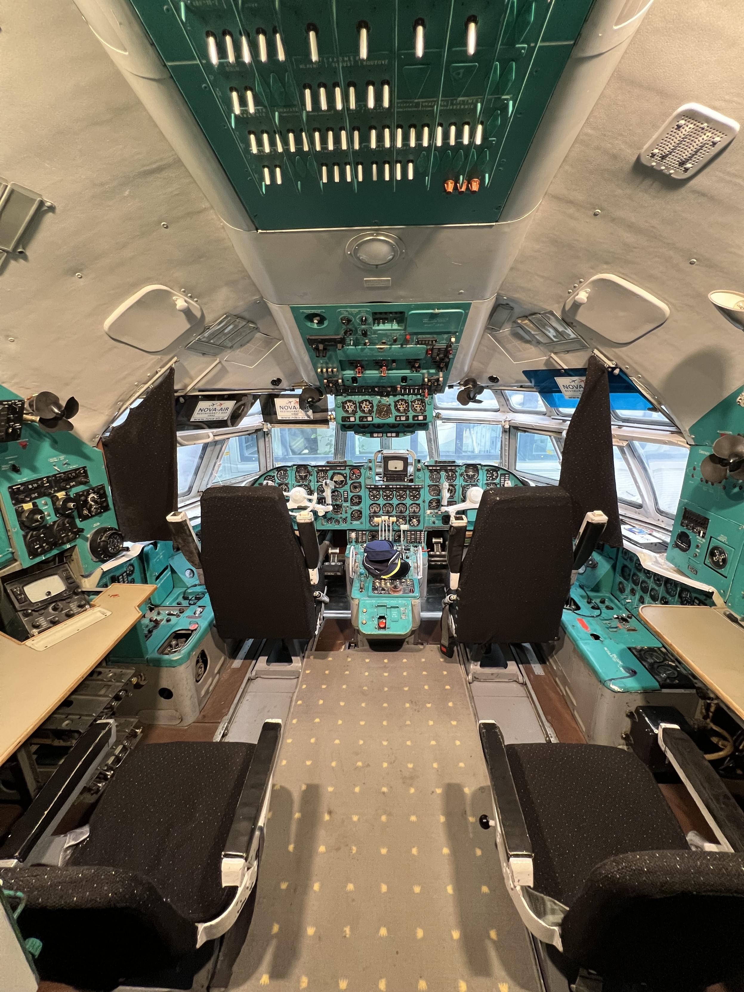 Cockpit eines Flugzeugs mit mehreren Sitzplätzen, zahlreichen Instrumententafeln und Schaltern an Decke und Wänden.