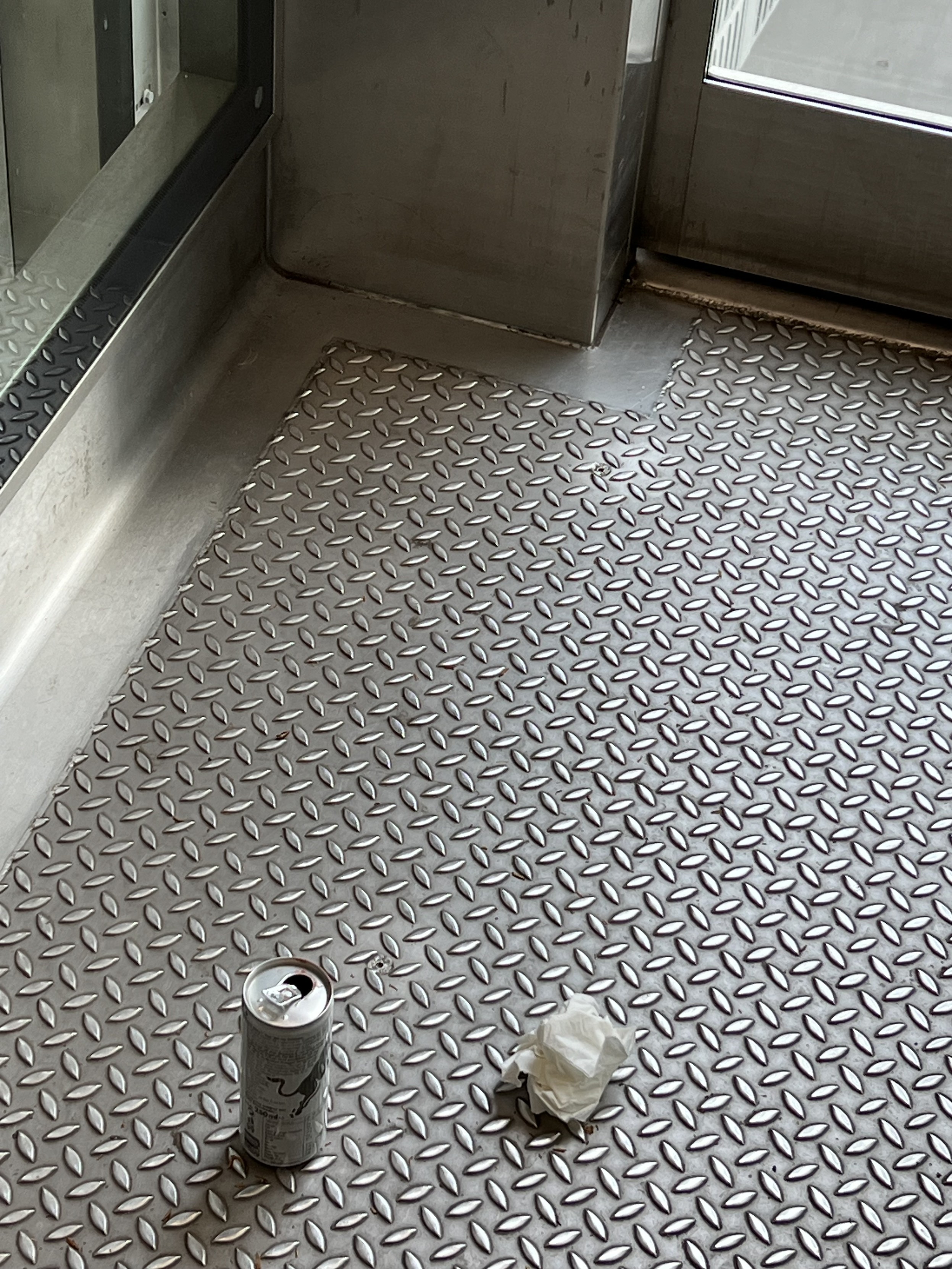 Am Boden eines Aufzugs steht eine geöffnete Getränkedose. Daneben liegt ein zerknülltes Papiertaschentuch. 