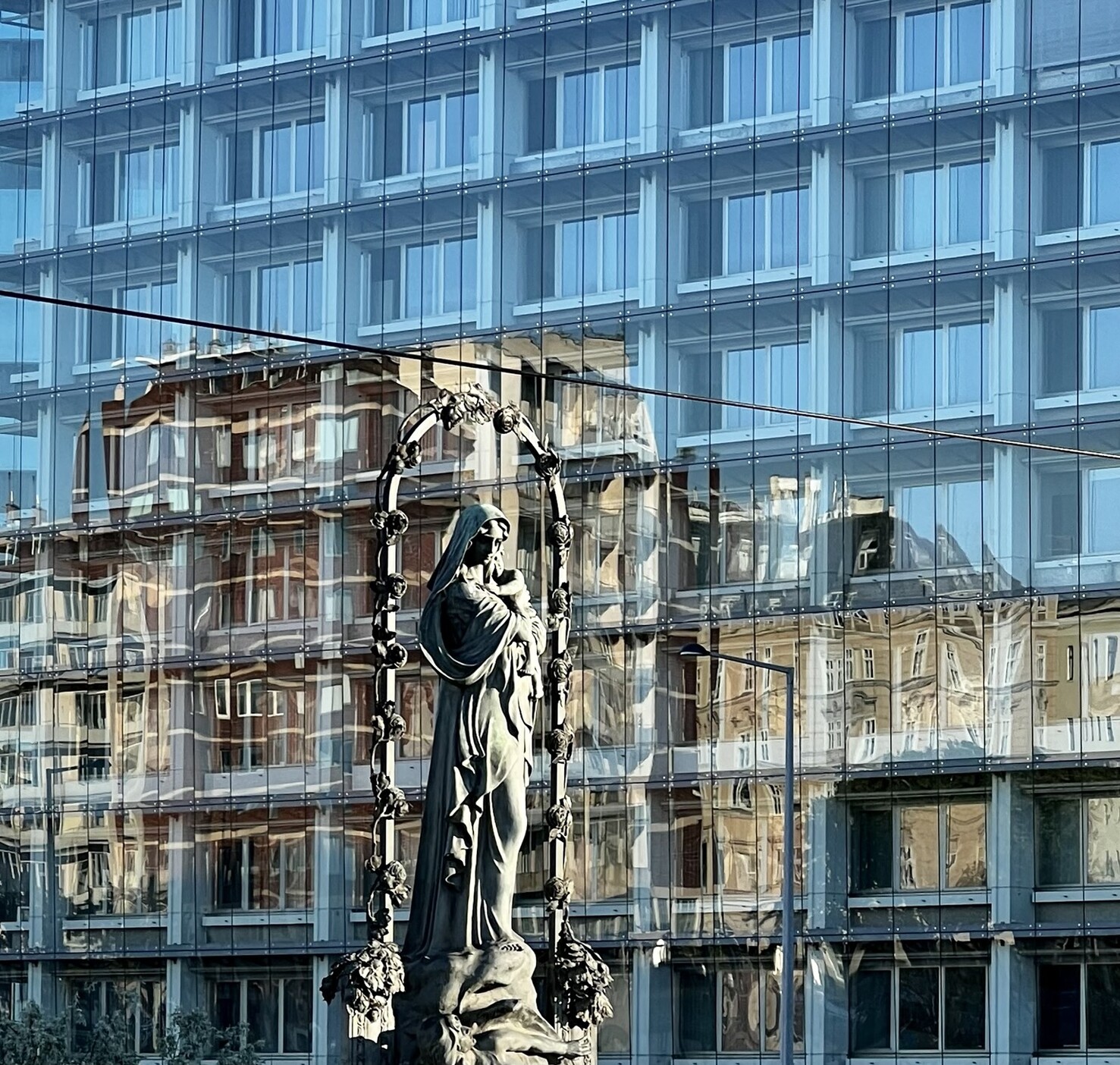 Marienstatue. Dahinter Gebäude mit Glasfront in der sich älteres Gebäude spiegelt.