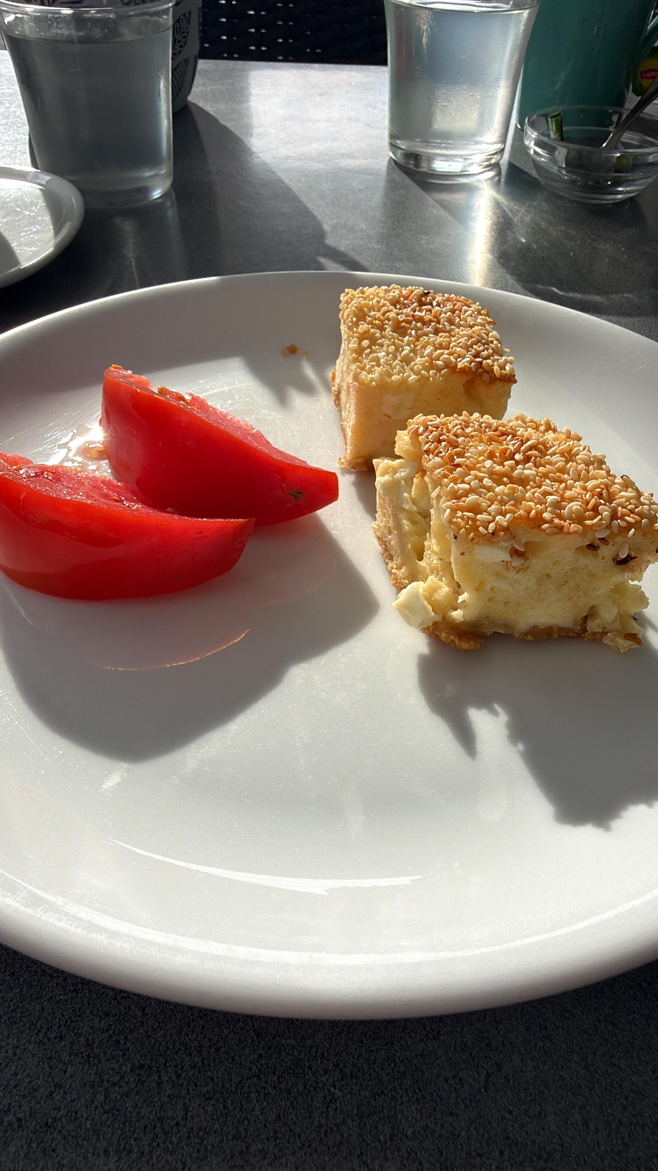 Weißer Teller mit zwei Gebäckstücken mit Sesam, zwei Tomatenscheiben. Zwei Gläser Wasser und Besteck auf einen sonnenbestrahlten Tisch auf dem die Dinge klare Schatten werfen.