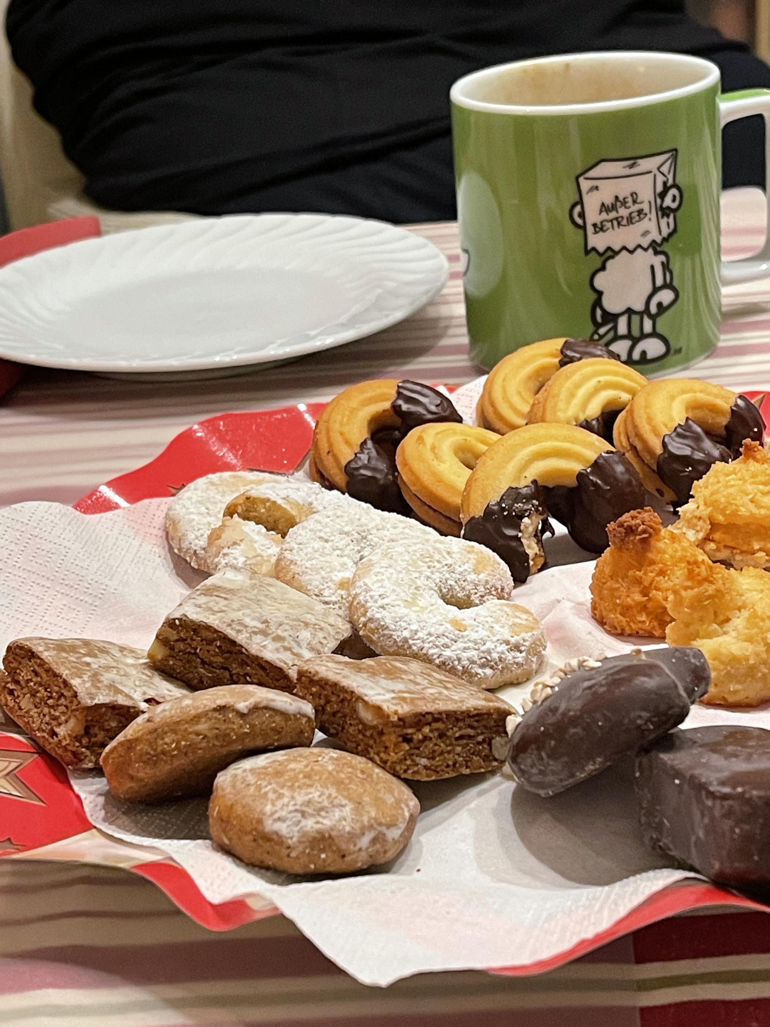 Ein Teller mit verschiedenen Weihnachtsplätzchen, darunter Lebkuchen, Vanillekipferl, Schoko-Kringel und Kokosmakronen, arrangiert auf einer roten Serviette. Im Hintergrund stehen ein leerer weißer Teller und eine grüne Tasse mit einer Comic-Figur und der Aufschrift „Außer Betrieb“.