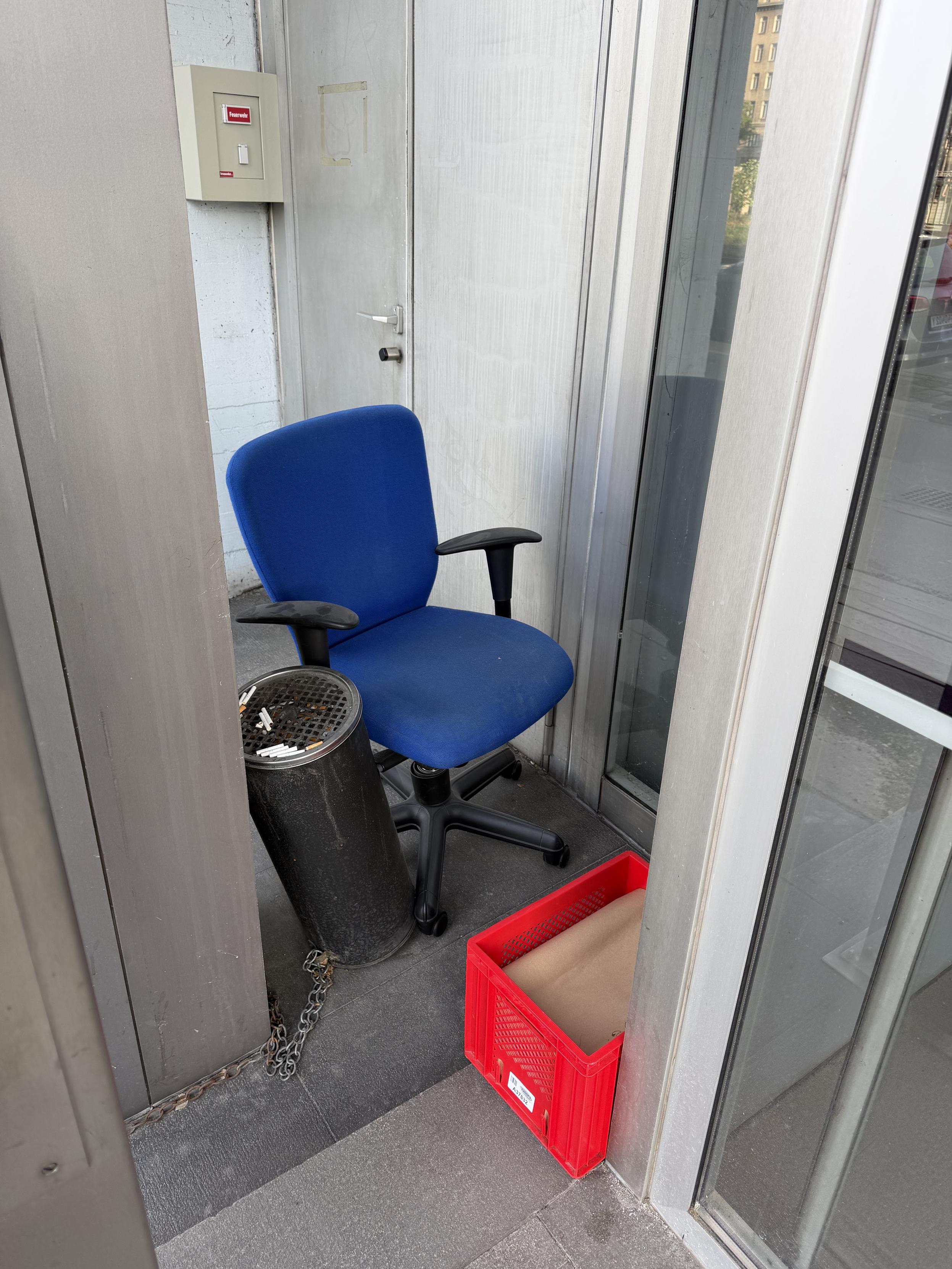 Blauer Bürostuhl in einer kleinen Nische neben einem Aschenbecher und einer roten Kiste, umgeben von grauen Wänden und Glasflächen.