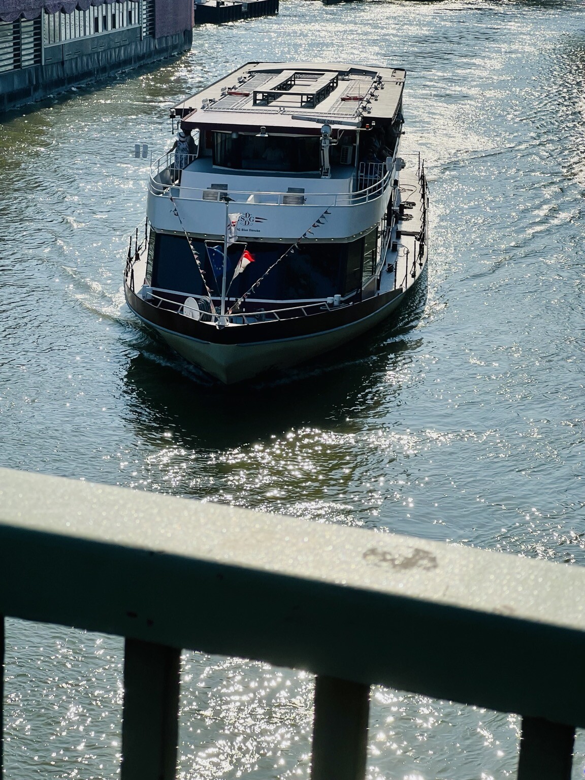 Brückengeländer. Dahinter Wasserstrom auf dem ein Schiff auf glitzernden Wasser fährt.