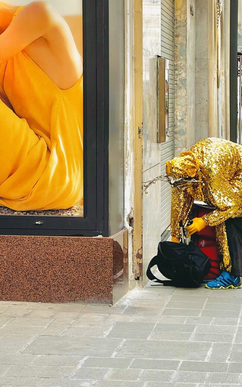 Straße. Links ein Werbeplakat mit einer sitzenden Frau in einem orange-goldenen Kleid. Rechts ein gebückter Mensch in einem Ganz-Körper-Gewand aus Gols und einem Art Geweih im Kopf. Sie kramt in einem Rucksack. 